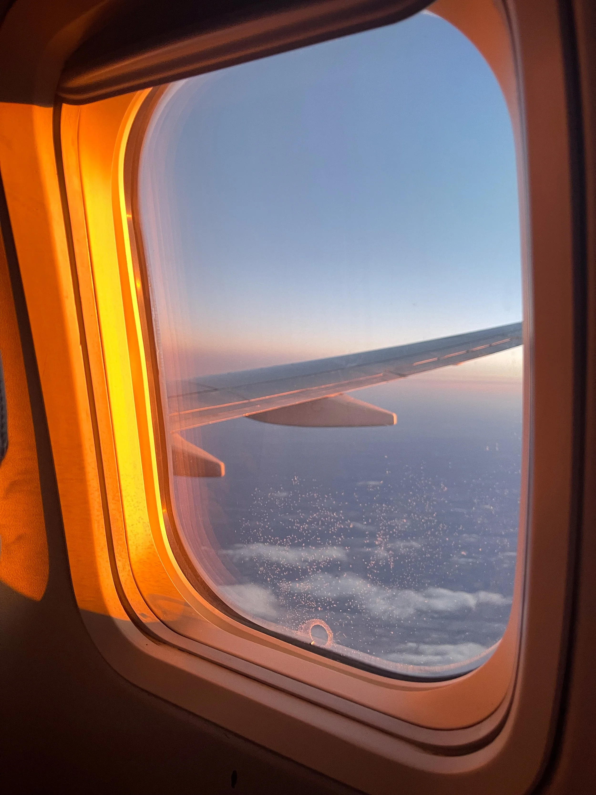 View from an airplane window showing the wing, clouds below, and a sky with a gradient of blue and pink at sunrise or sunset.