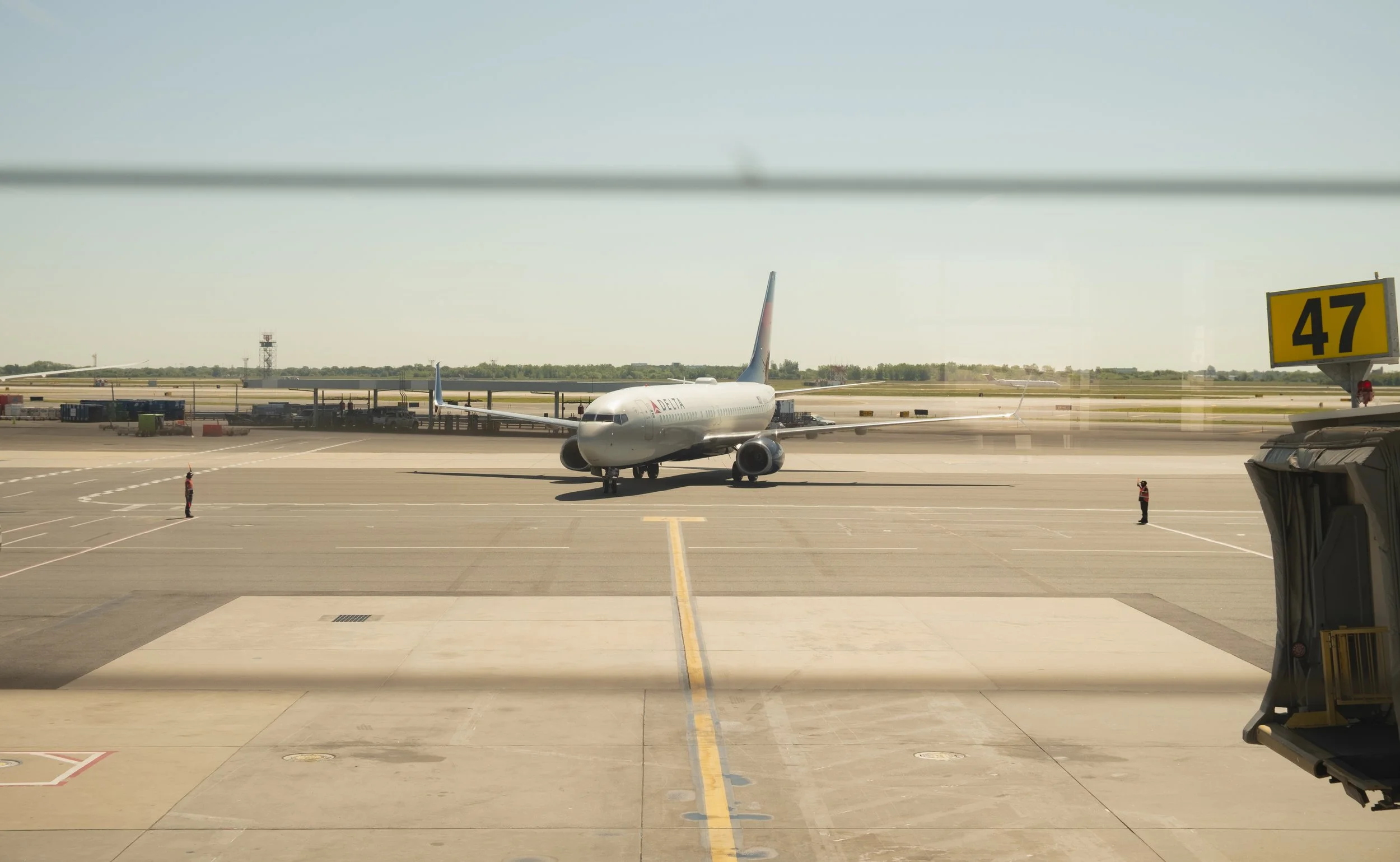 Airplane parked on the tarmac at an airport, viewed from inside a terminal through a window. The airplane is white with Delta branding and is positioned near gate 47.