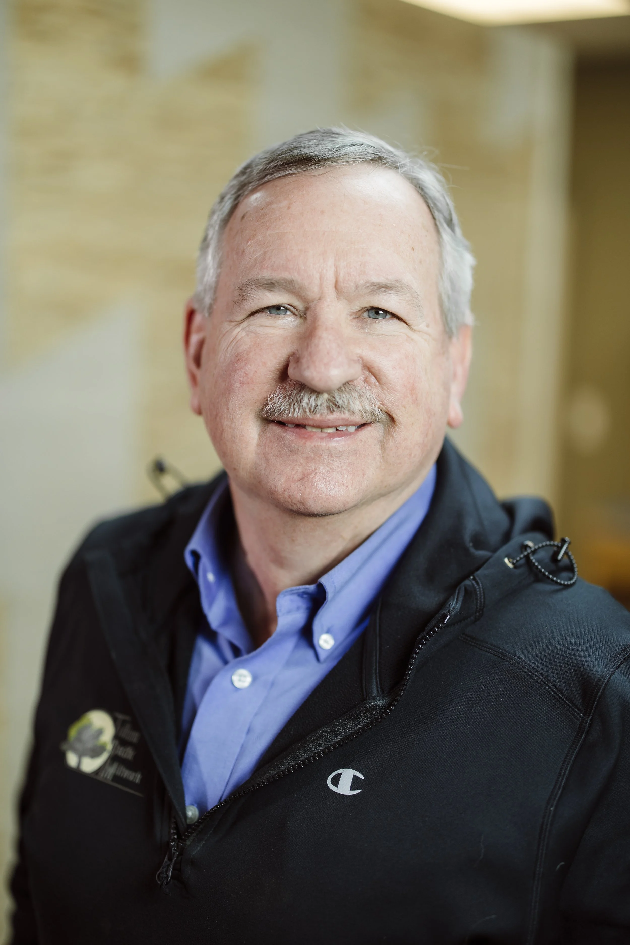 A portrait of a smiling middle-aged man with gray hair, wearing a dark blazer and blue shirt, against a neutral background.
