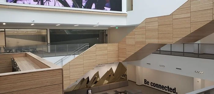 Modern shopping mall interior with wooden staircase and escalator, with a partial view of an upper level and a large digital screen.