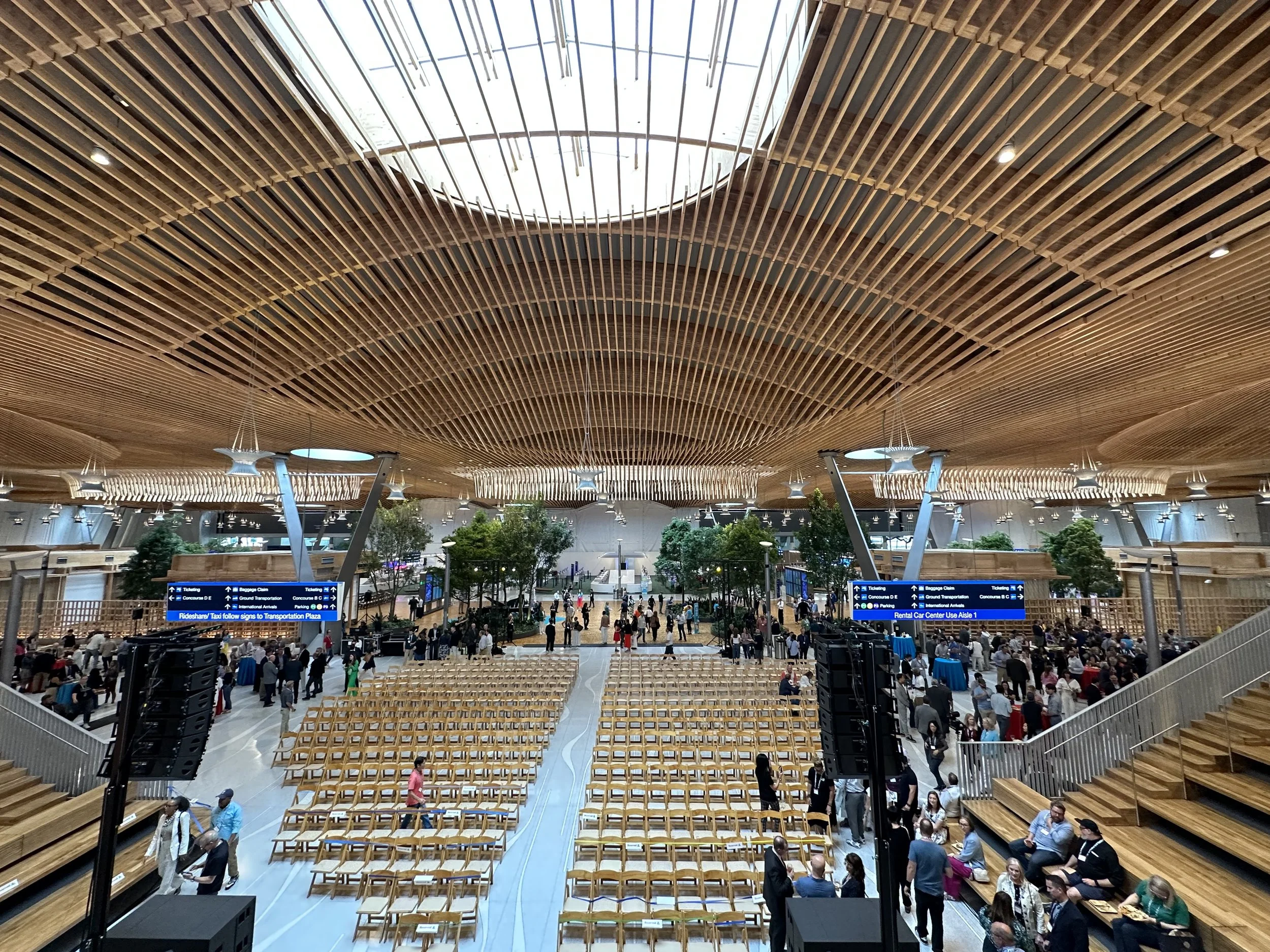 Inside a modern transportation hub or airport with a high wooden ceiling and a wide open space filled with seating, trees, and many people walking or gathering. Digital signs hang across the space displaying transportation information.