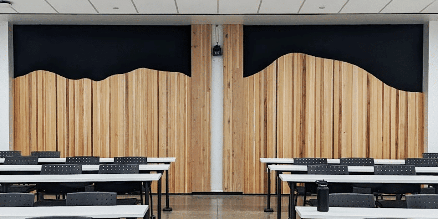Empty classroom with black chairs and white tables, and a wooden panel wall at the front of the room.