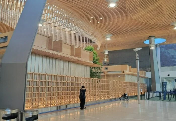 Interior of a modern building with wooden architectural elements, greenery, and few people; includes a person walking, a bicycle, and seating areas.