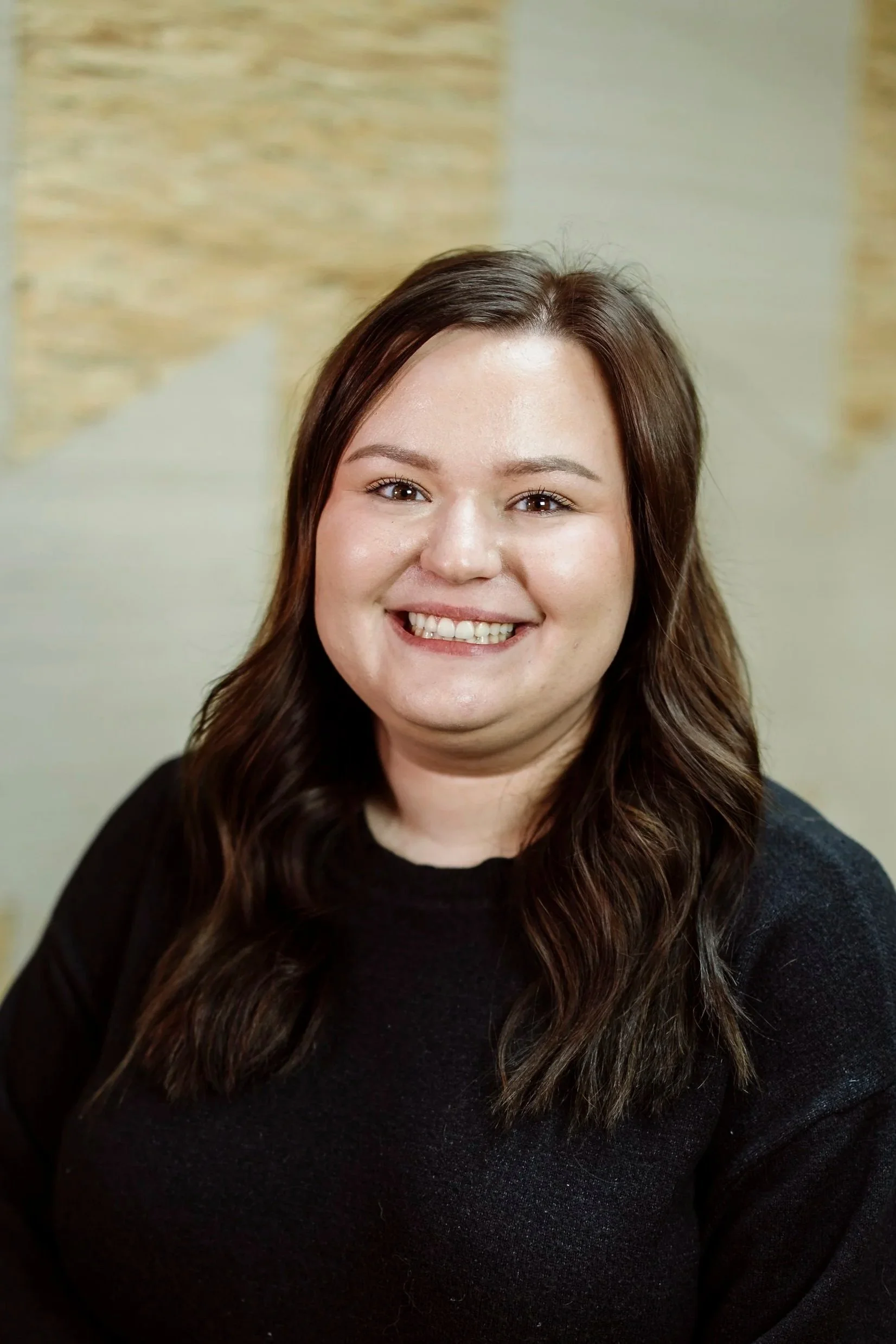 A woman with shoulder-length dark hair and a broad smile, wearing a black top, standing against a plain, light-colored background.