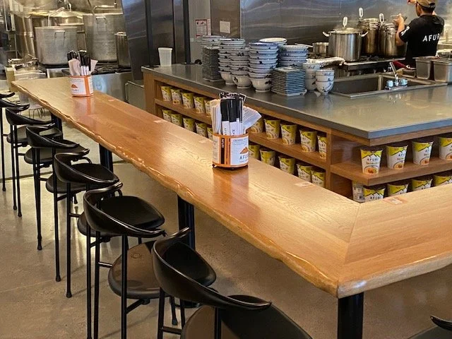Empty restaurant counter with black chairs, bowls, and containers, with a kitchen in the background.