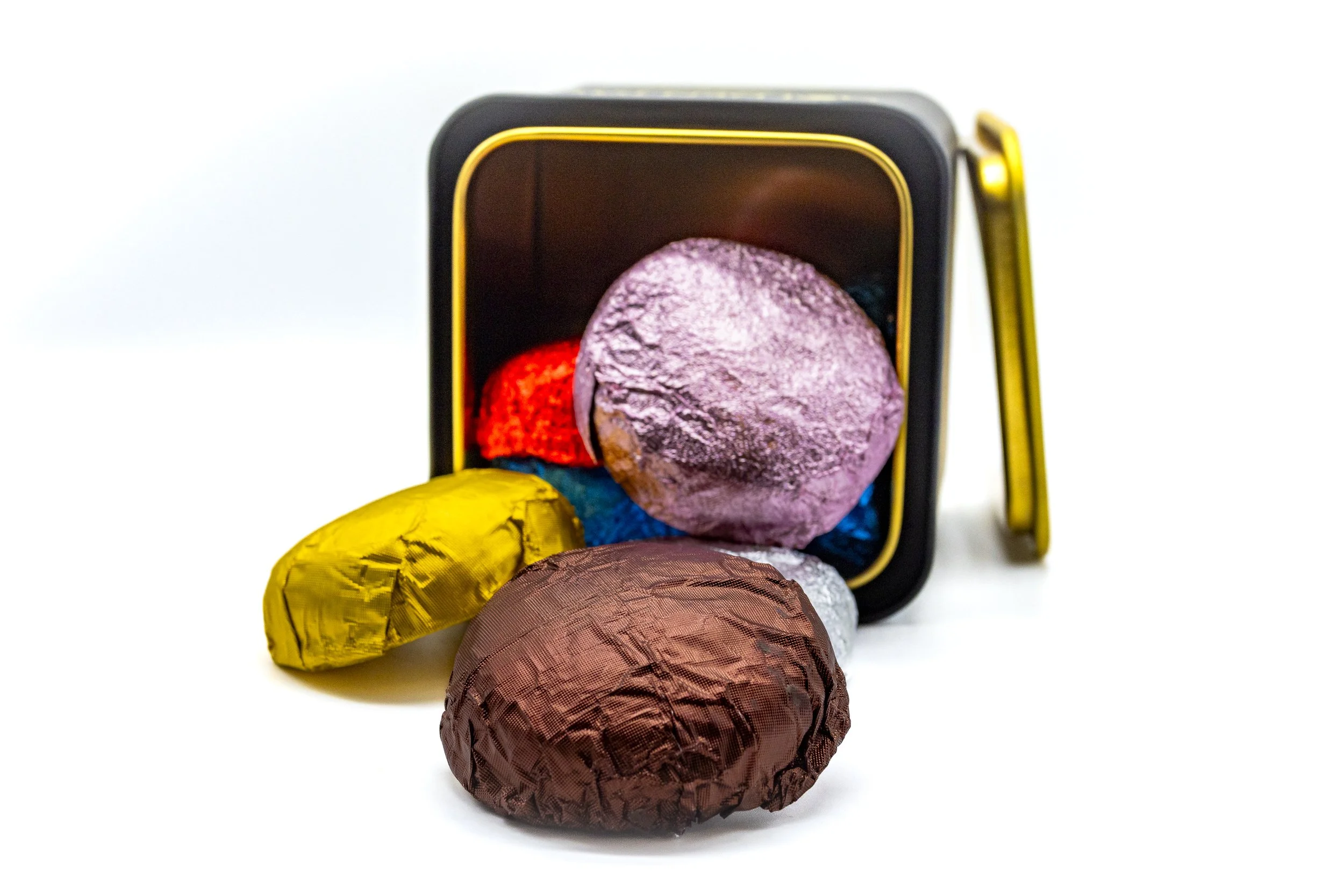 Colorful bath bombs inside and outside a black and gold box, with some spilling out onto a white surface.