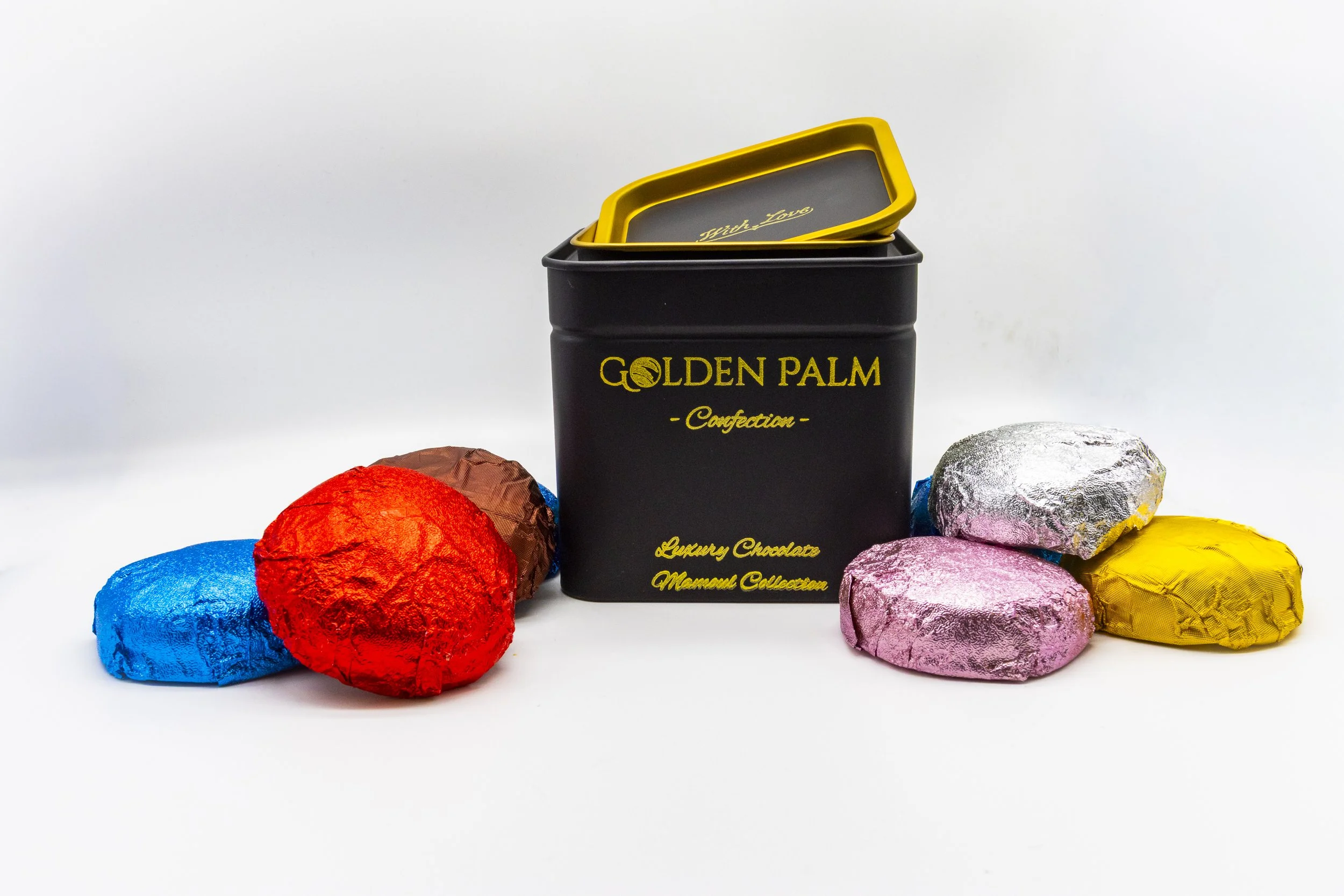 A black tin container with yellow text reading 'GOLDEN PALM Confection' and 'Luxury Chocolate Mamon Collection,' surrounded by colorful wrapped chocolates in blue, red, brown, pink, silver, and yellow foil, on a white background.