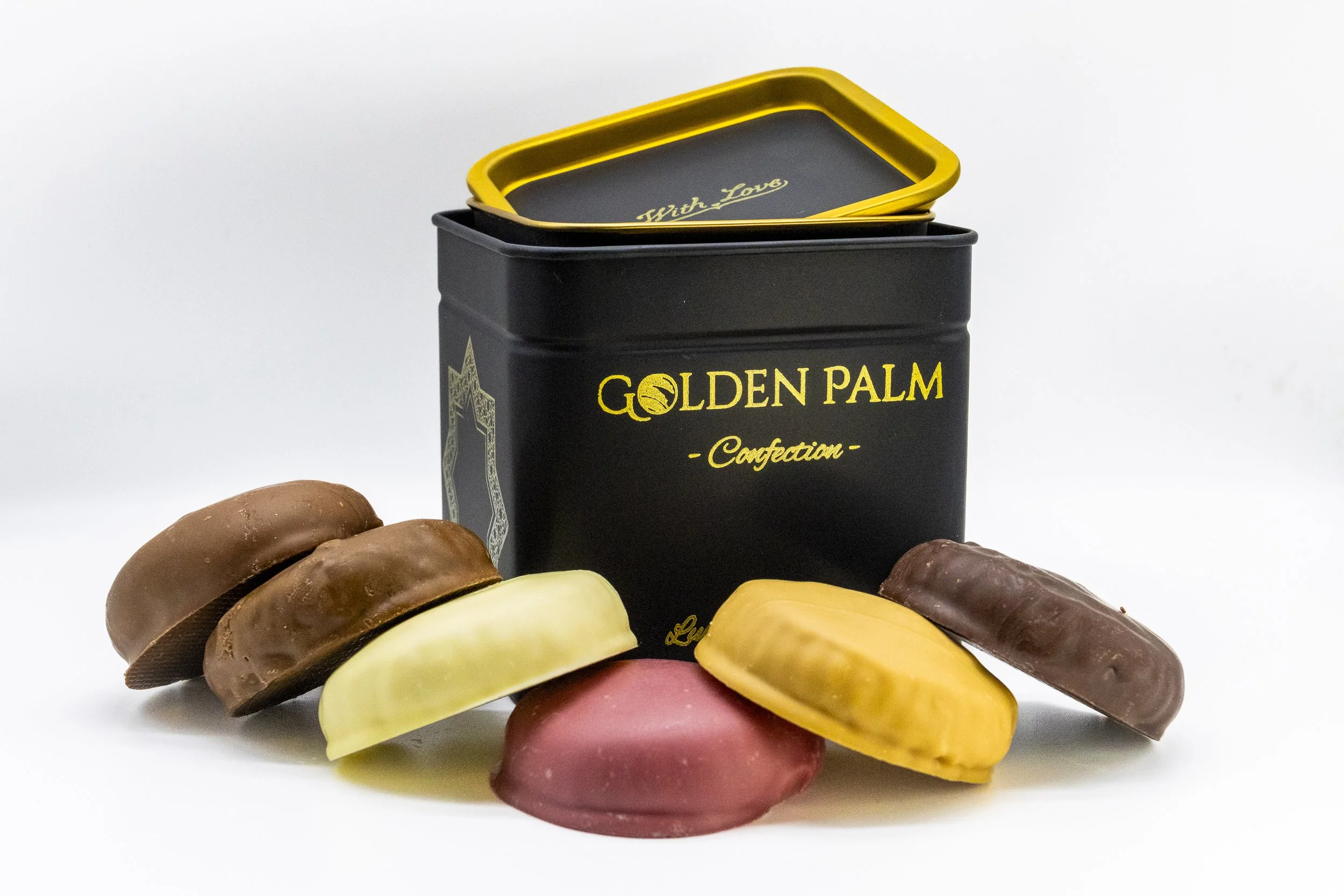 Assorted chocolate candies surrounding a black tin labeled 'GOLDEN PALM Confection' with an open gold-trimmed lid on a white background.