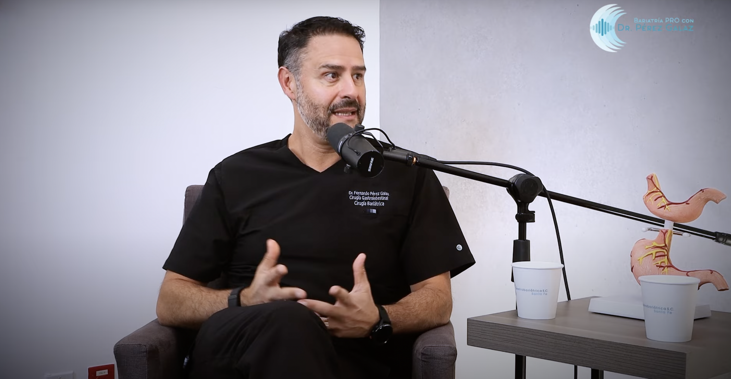 Un hombre con barba y cabello oscuro hablando en un podcast o entrevista, usando ropa médica negra, sentado en una silla y gesticulando con las manos, con micrófono frente a él y un modelo anatómico del estómago y los intestinos en una mesa al lado, en un ambiente de consulta médica.