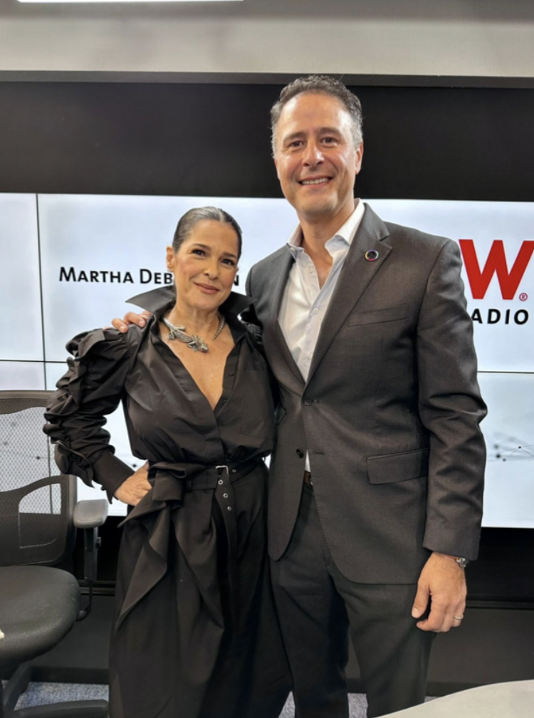 Dos personas, una mujer y un hombre, posando juntos en un estudio de radio, con un cartel que dice 'Martha Debayle' y 'W Radio' en el fondo.