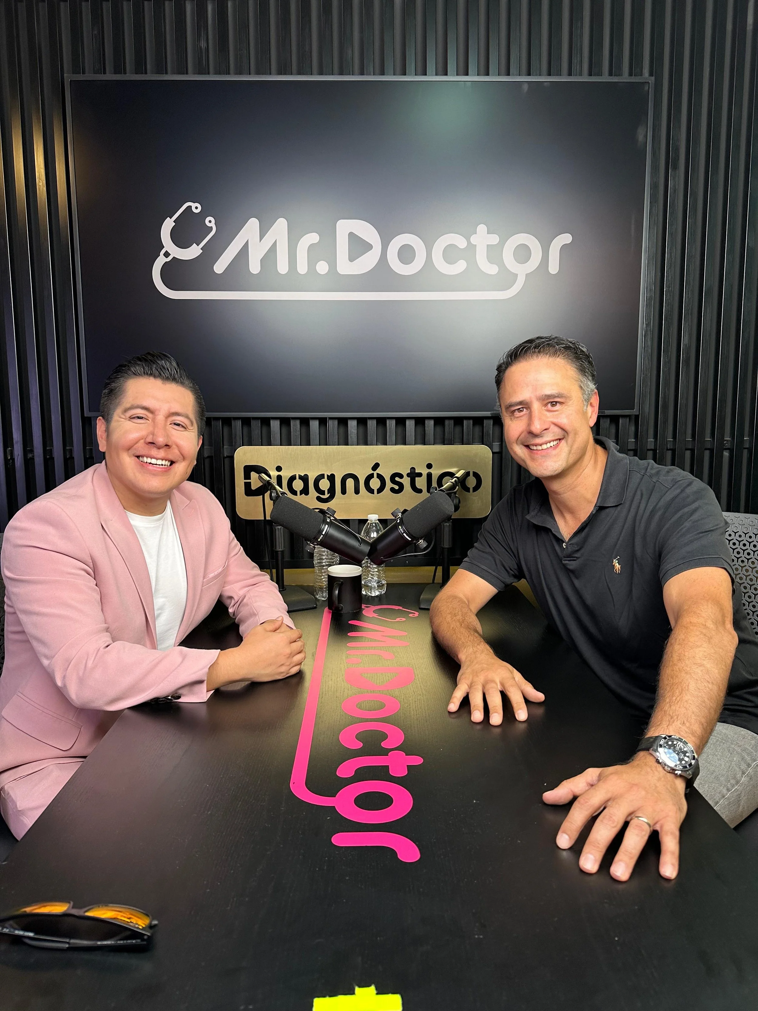 Dos hombres sonriendo y sentados en una mesa durante una grabación, con micrófonos frente a ellos y un cartel que dice 'diagnóstico' detrás, en un estudio con fondo negro y logotipo que dice 'Mr. Doctor' con un estetoscopio en el diseño.