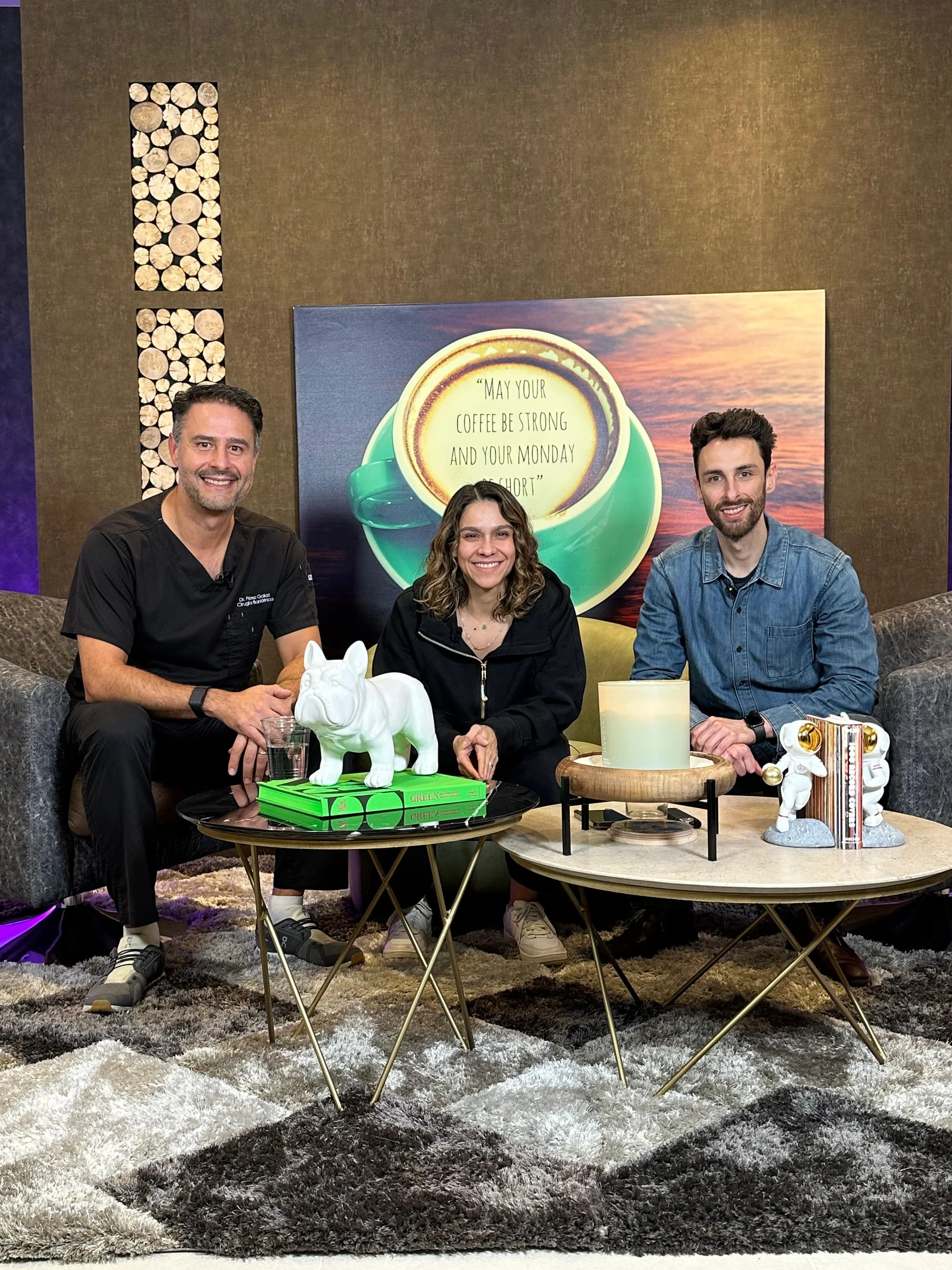Tres personas sentadas en un sofá, dos hombres y una mujer, rodeados de mesas con objetos decorativos, una imagen de una taza con la frase "May your coffee be strong and your Monday short" al fondo.
