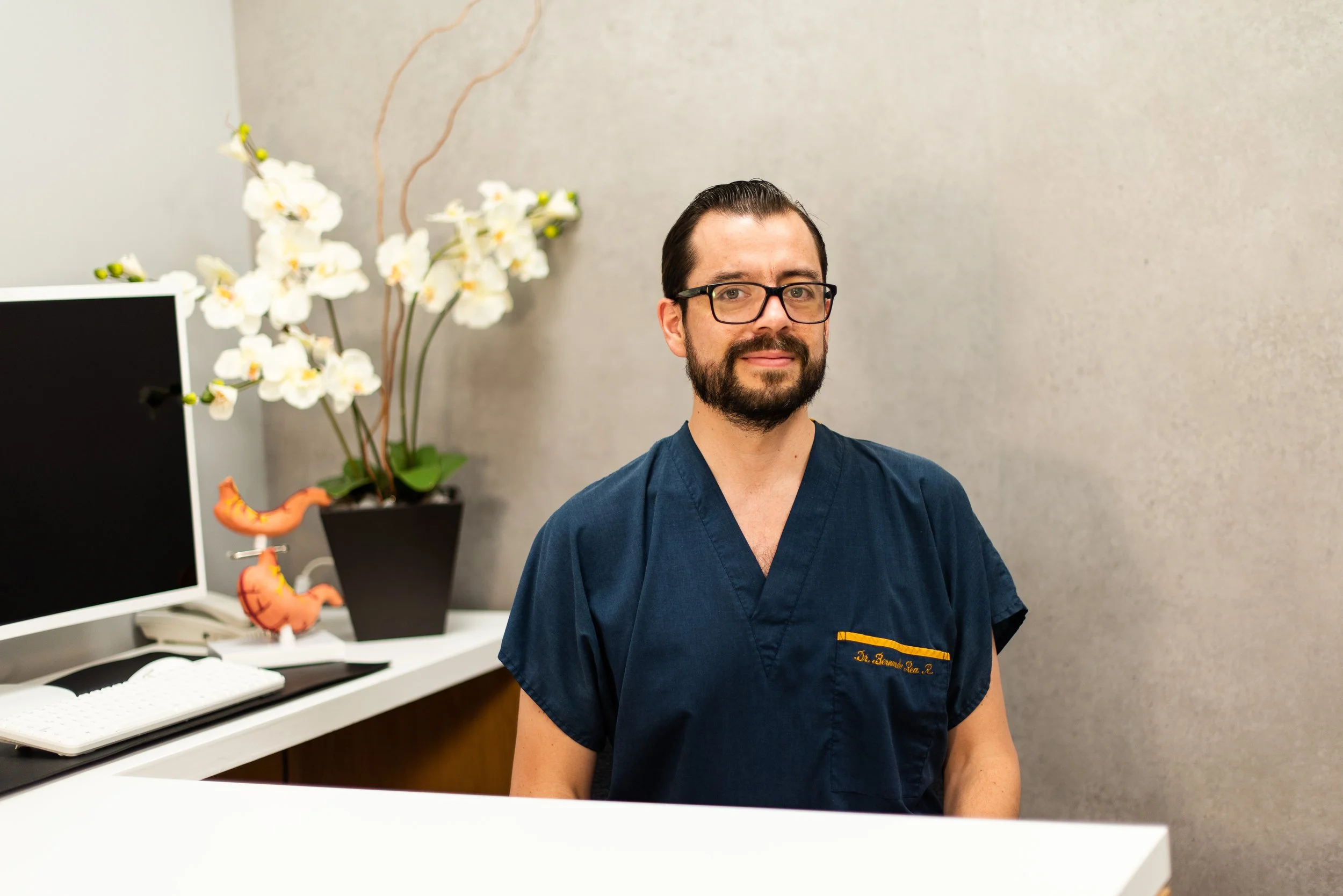 Un hombre con barba y gafas, vestido con uniforme médico azul, sentado en una recepción en un consultorio con plantas y decoraciones en el fondo.