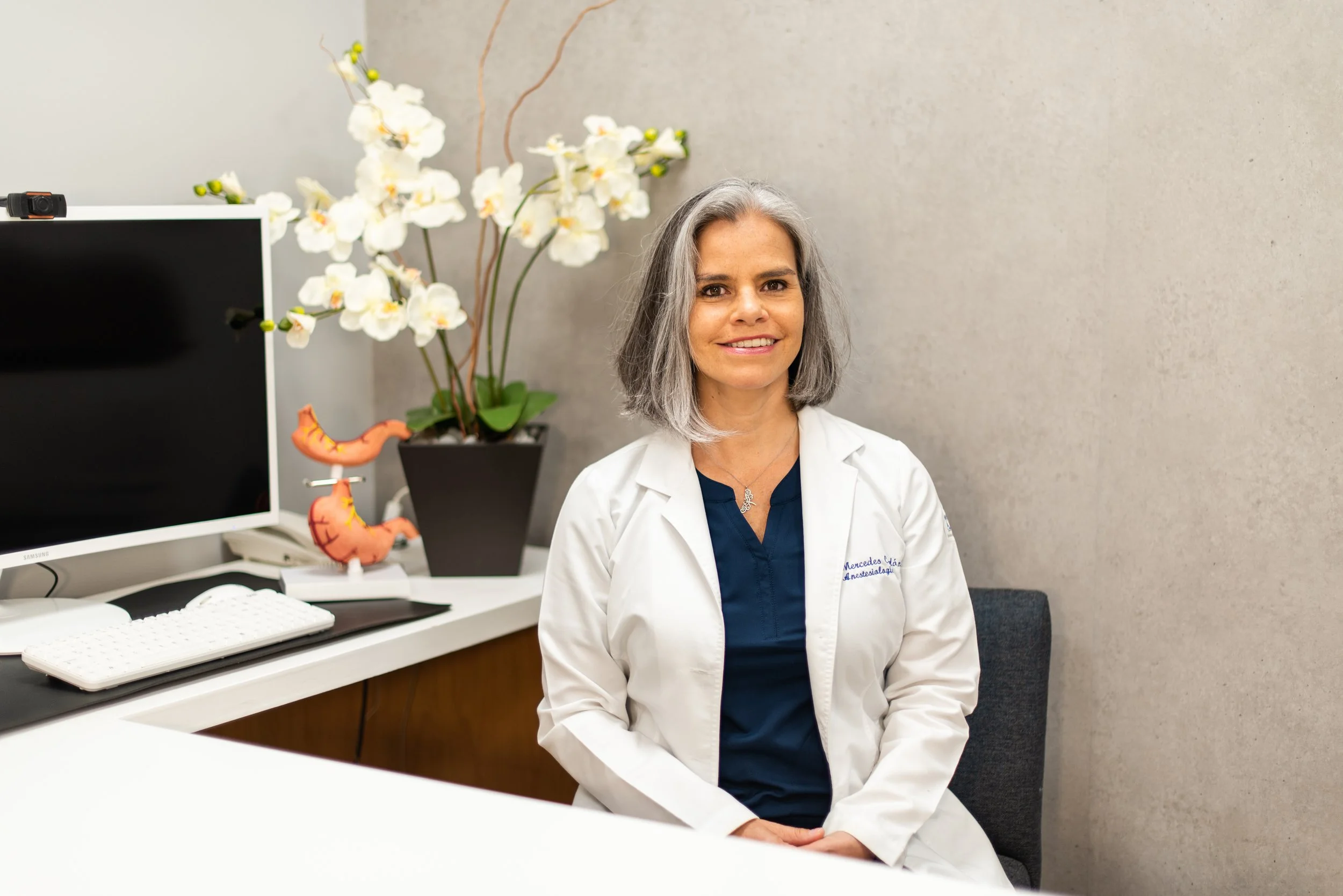 Mujer con cabello canoso, vestida con bata blanca y blusa azul, sentada en una oficina con computadora y plantas de orquídeas blancas de fondo.