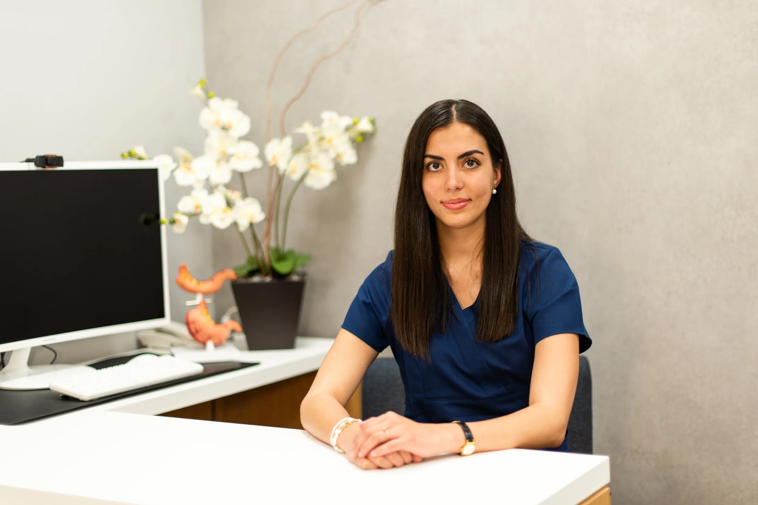 Mujer joven con cabello largo y oscuro, vestida con uniforme azul, sentada en un escritorio en una oficina, con una planta de flores blancas y decoraciones en el fondo.