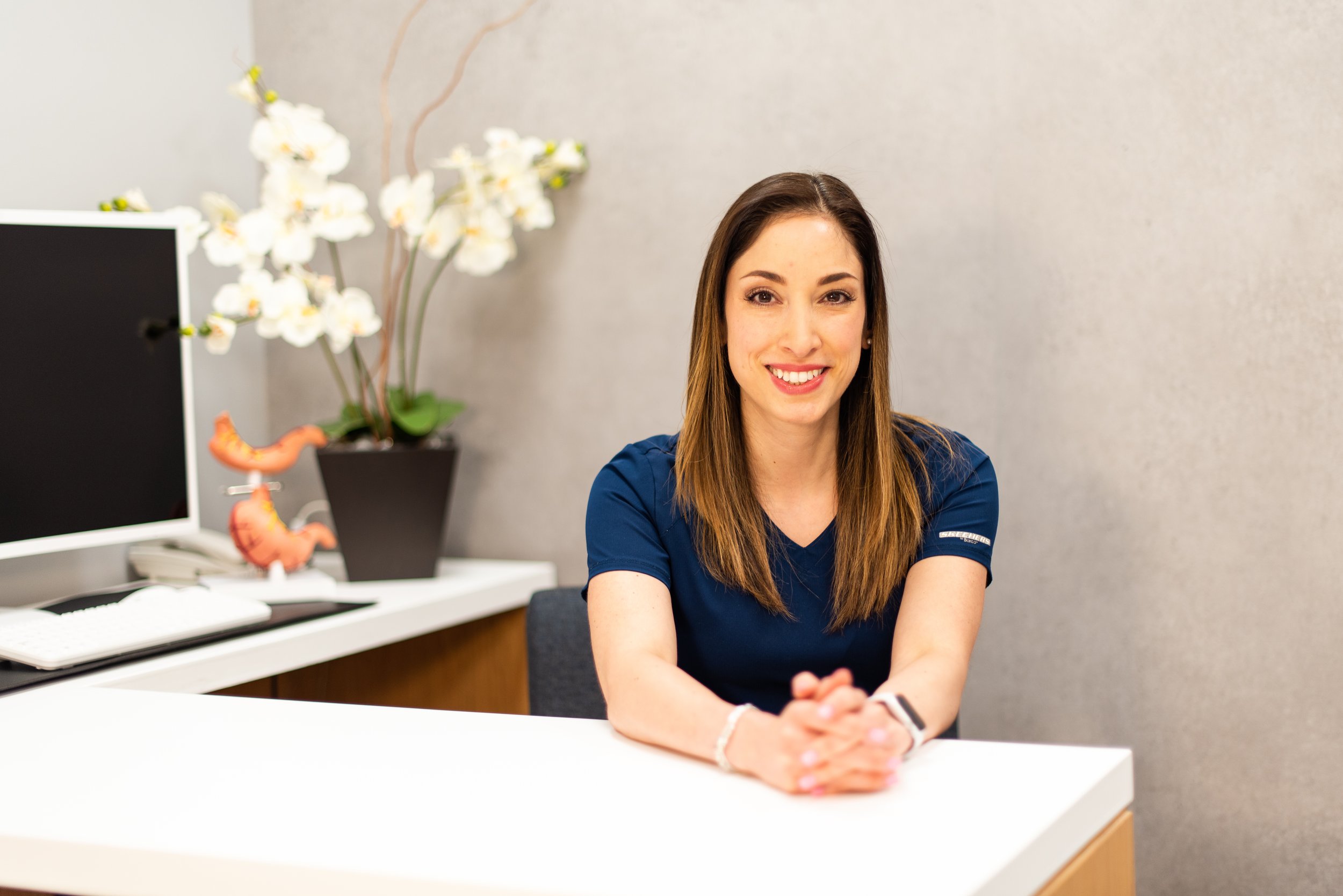 Mujer sonriendo en una oficina con plantas y un monitor en el fondo.