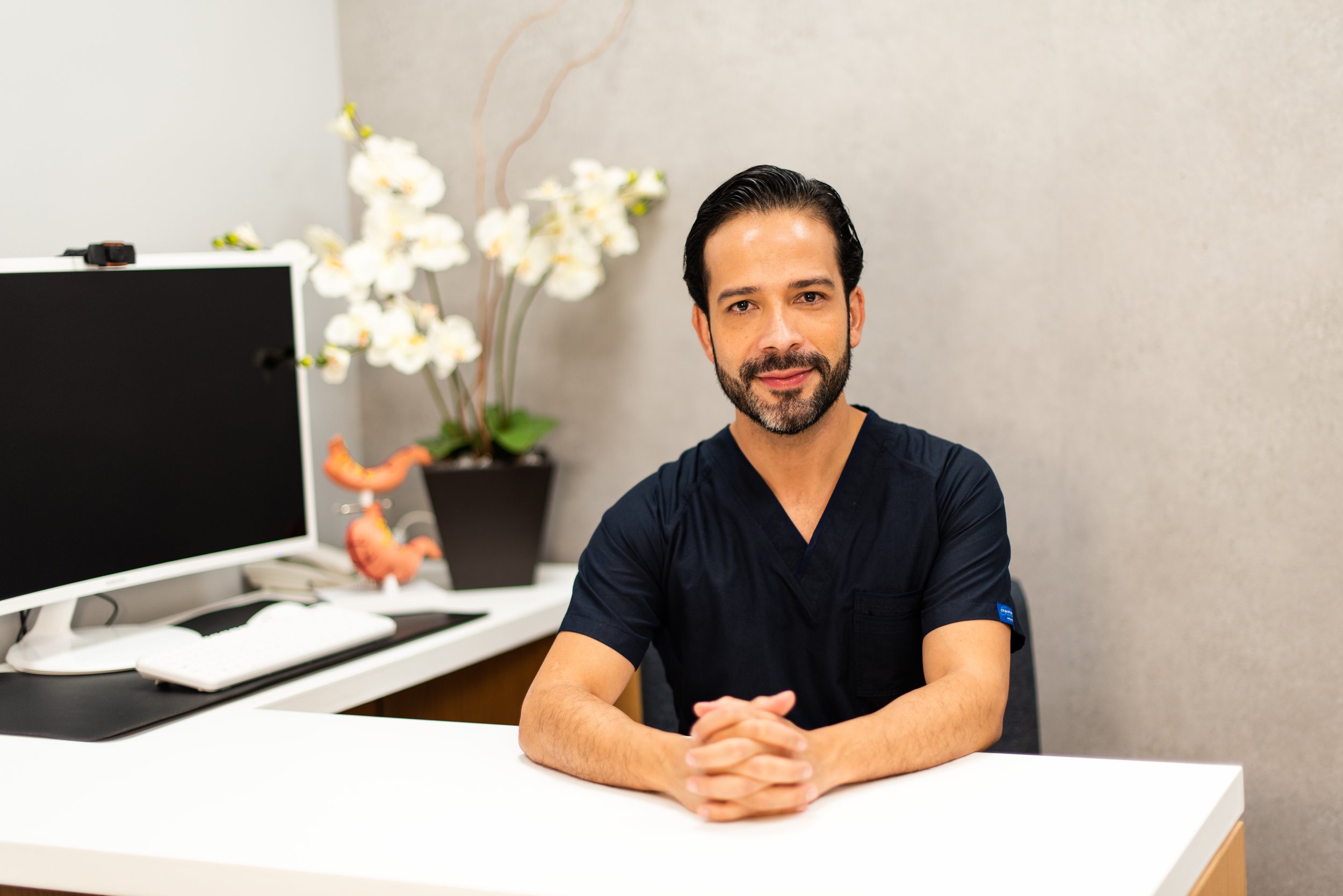Hombre con barba y cabello oscuro, sentado en un escritorio con las manos entrelazadas, en una oficina con fondo gris, plantas y una pantalla de computadora.