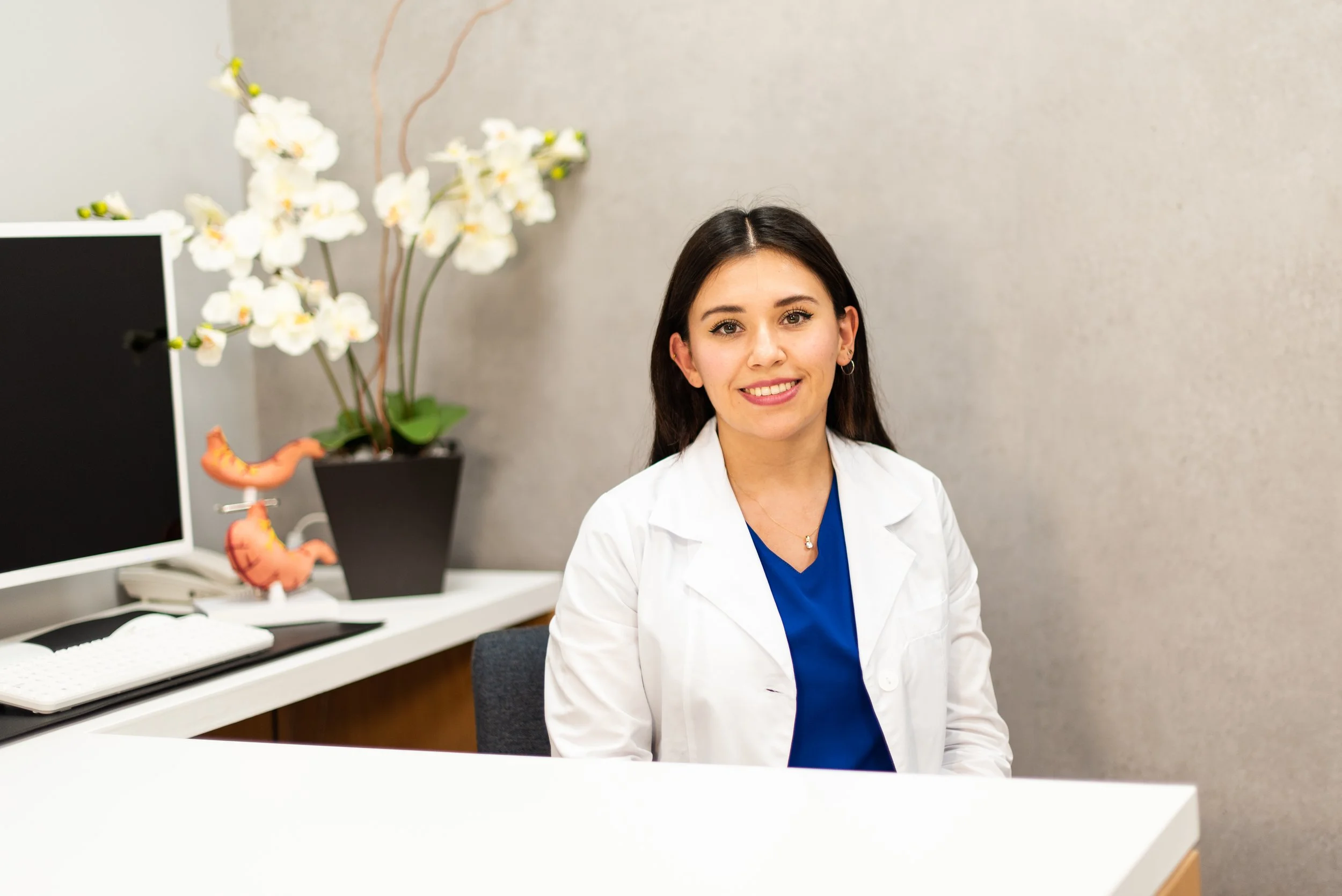 Mujer con bata blanca y blusa azul sentada en escritorio de oficina, con monitor, libro, plantas y modelos anatómicos de estómago en el fondo.