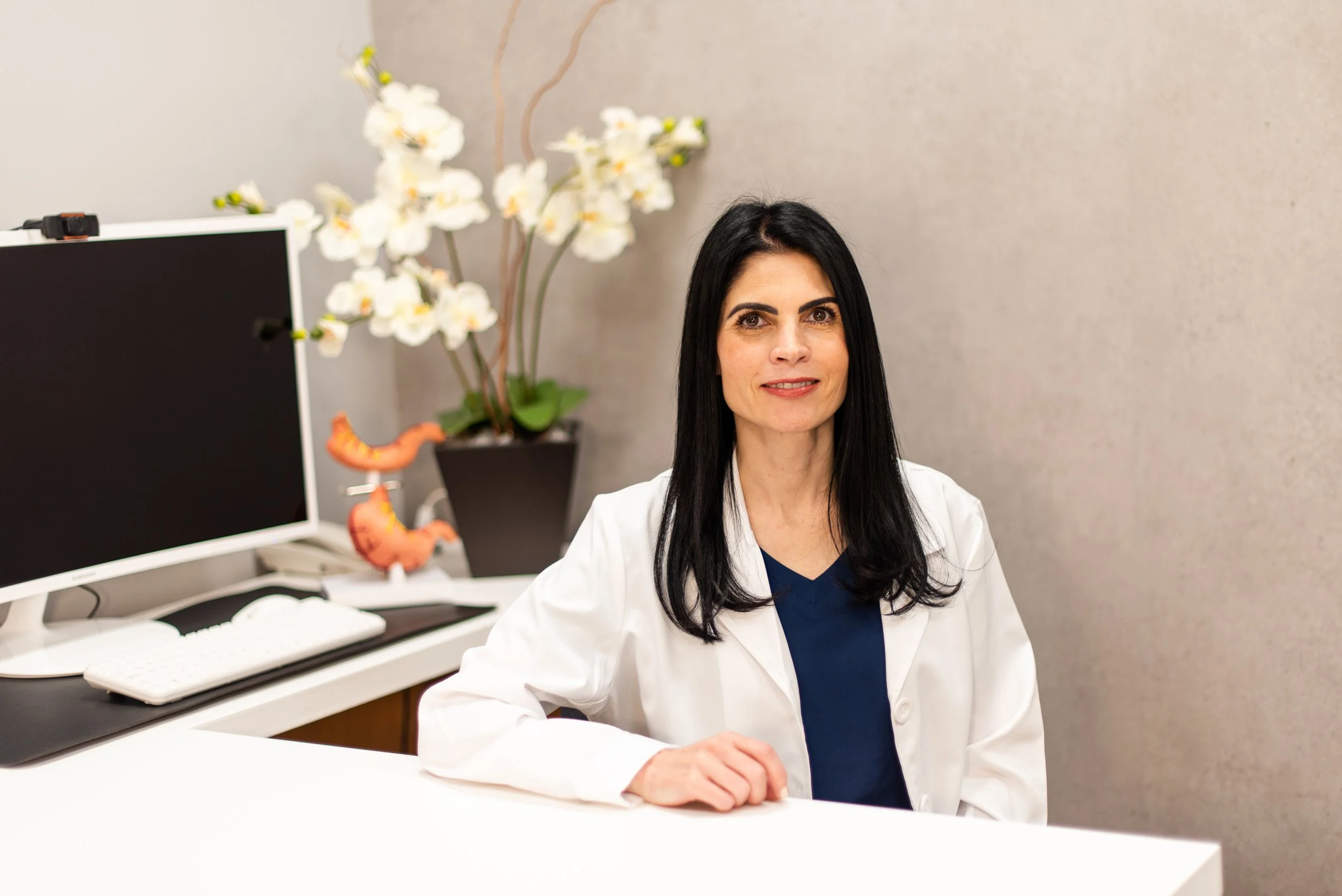 Mujer doctora en oficina con florero y monitor en el fondo.