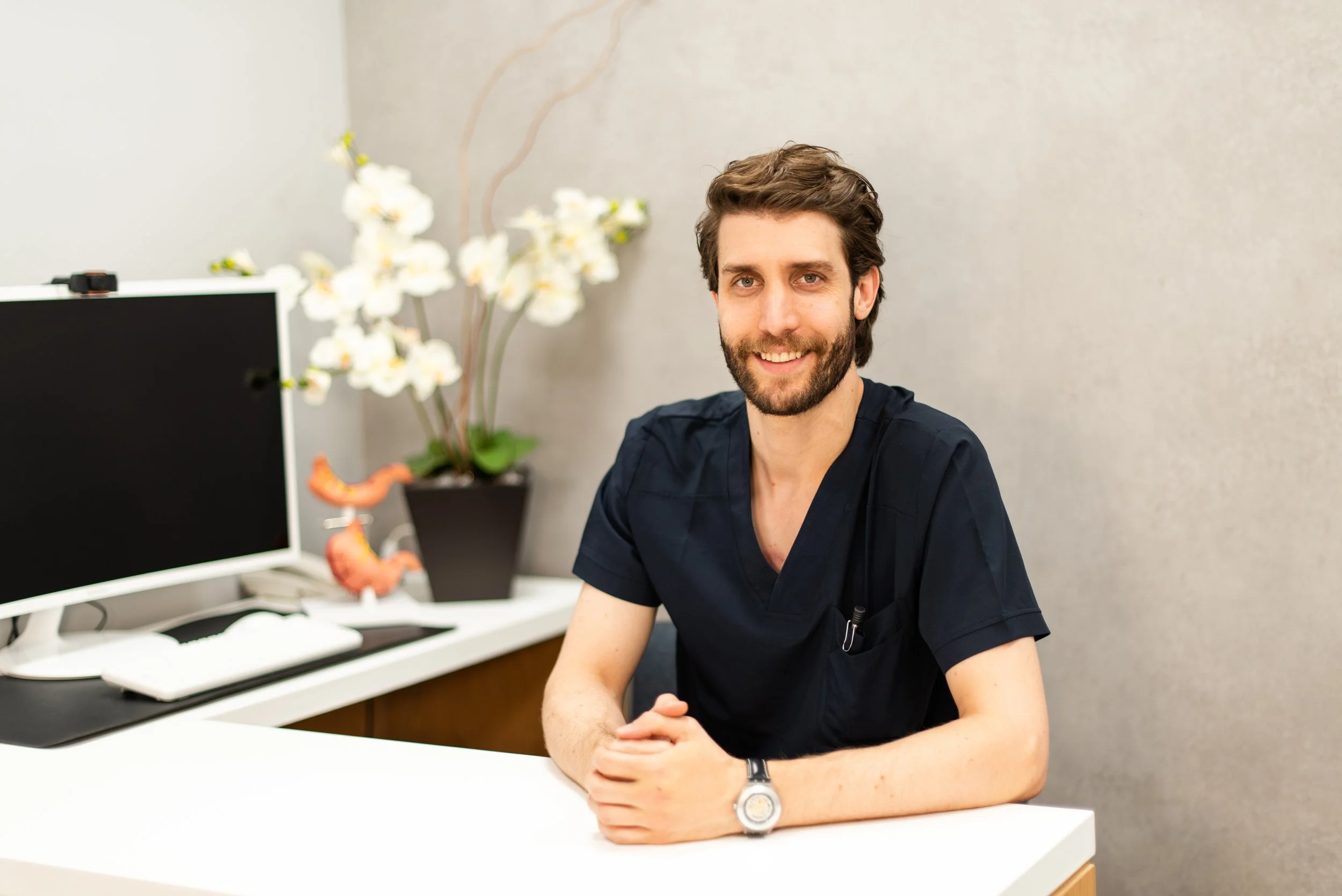 Un joven profesional de cabello castaño, barba y sonrisa, sentado en una oficina moderna con una computadora, una planta con flores blancas y una impresión de un órgano en el fondo.