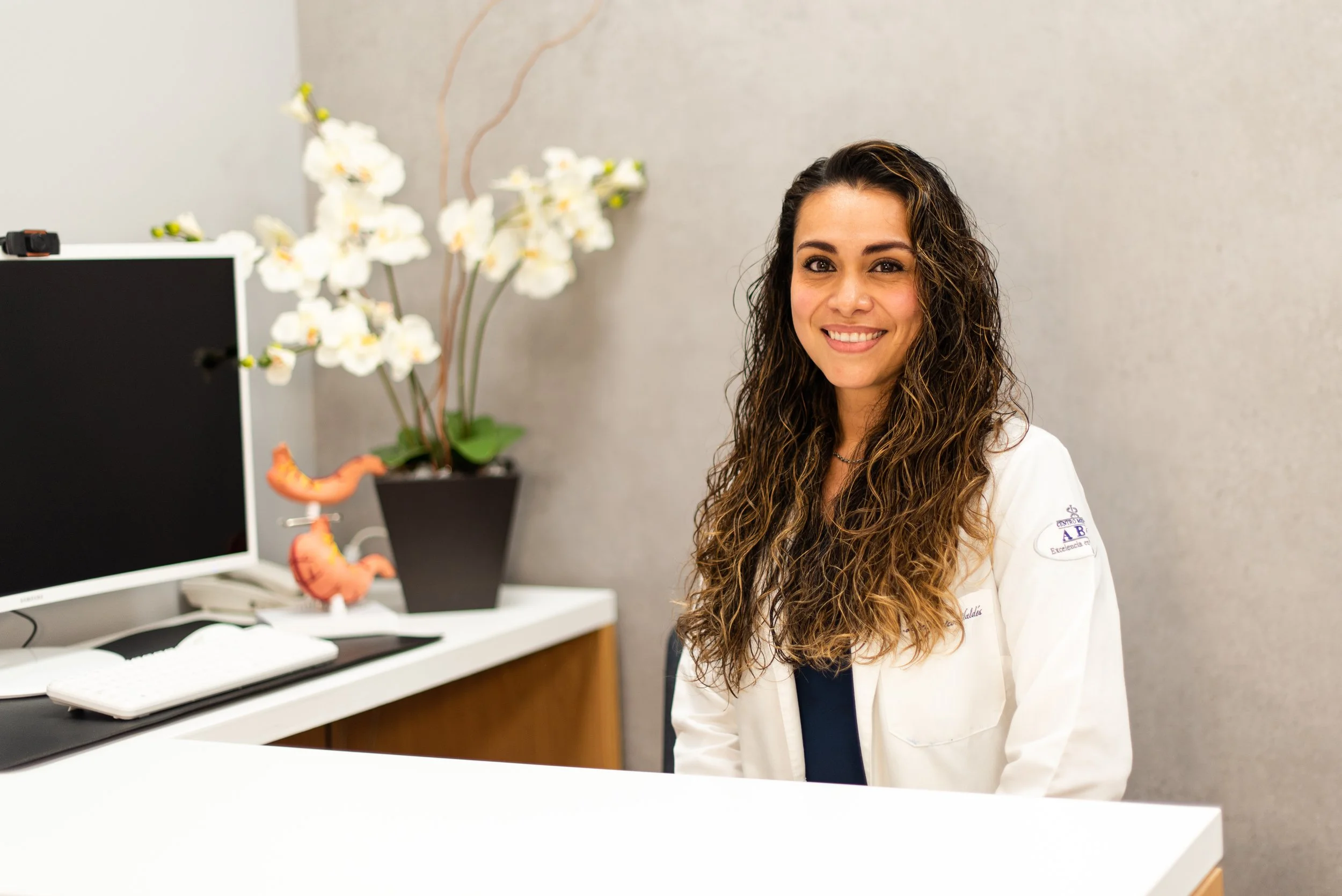 Mujer con cabello rizado en mostrador de oficina, con planta de orquídeas y pantalla de computadora en el fondo.