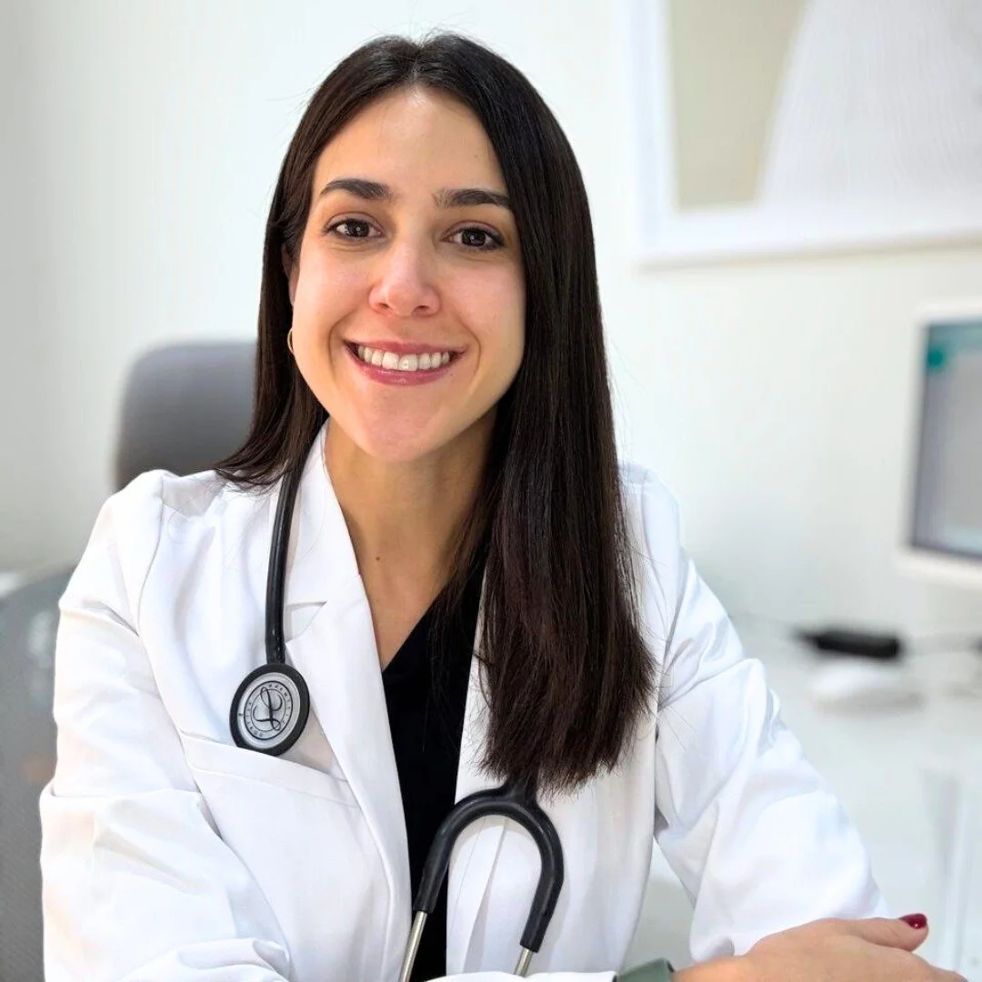 Mujer doctora sonriendo, con bata blanca y estetoscopio, en un consultorio médico.