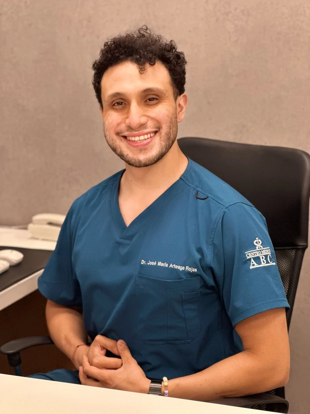Hombre sonriendo, vestido con uniforme de médico en una oficina.