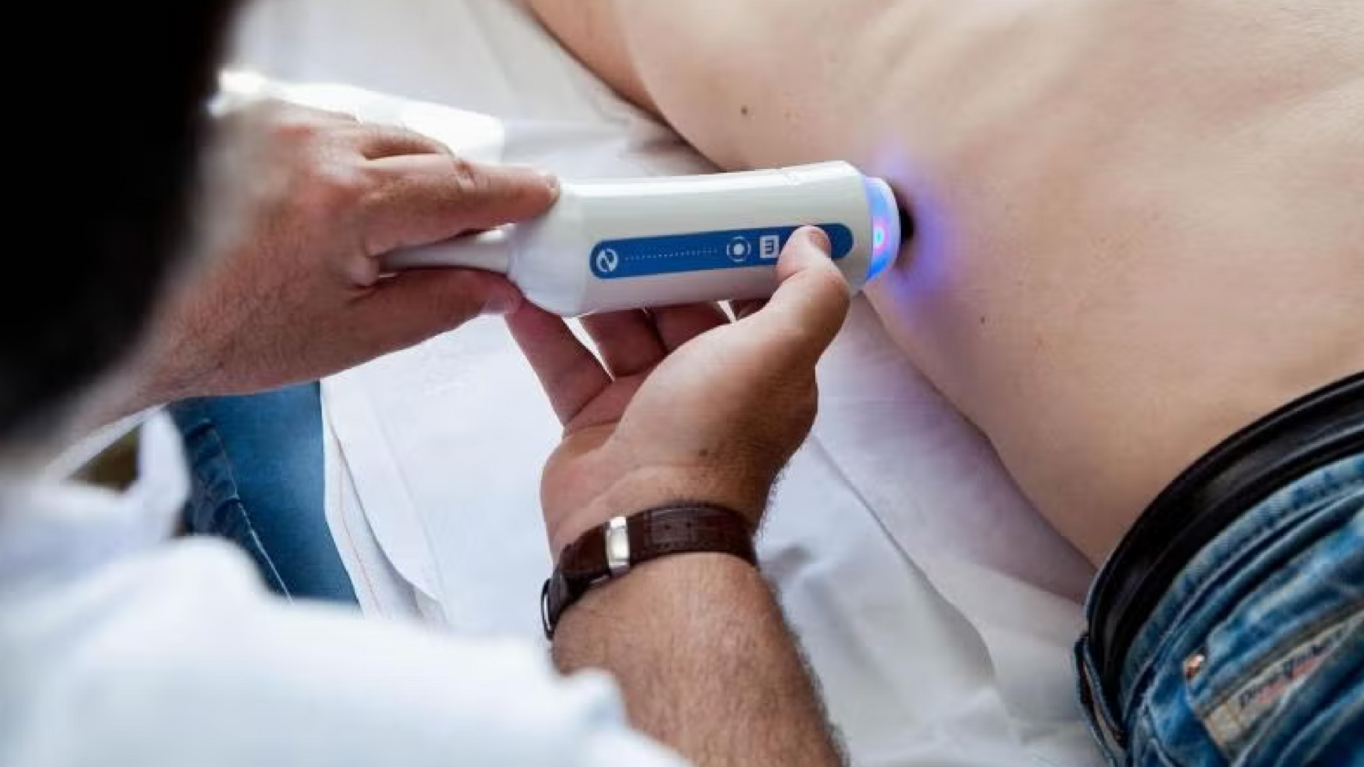 Médico realizando una tomografía o escaneo en la zona lumbar de la espalda de un paciente. La máquina tiene una luz azul y el paciente está acostado boca abajo.