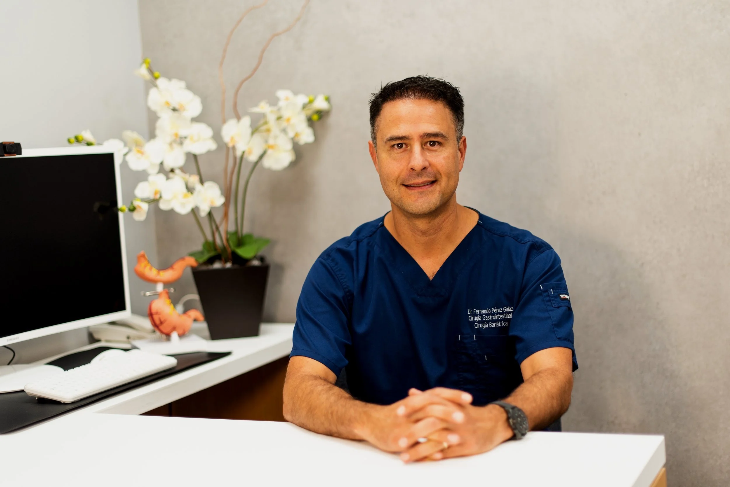 Un hombre con bata azul sentado en un escritorio, sonriendo, en un consultorio con plantas ornamentales y una pantalla de computadora