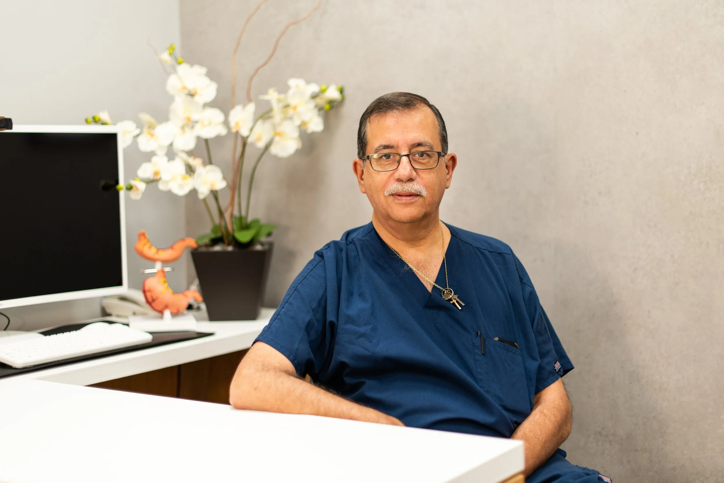 Un hombre con gafas y camisa azul sentado en una oficina, con una planta de orquídea y un monitor en el fondo.