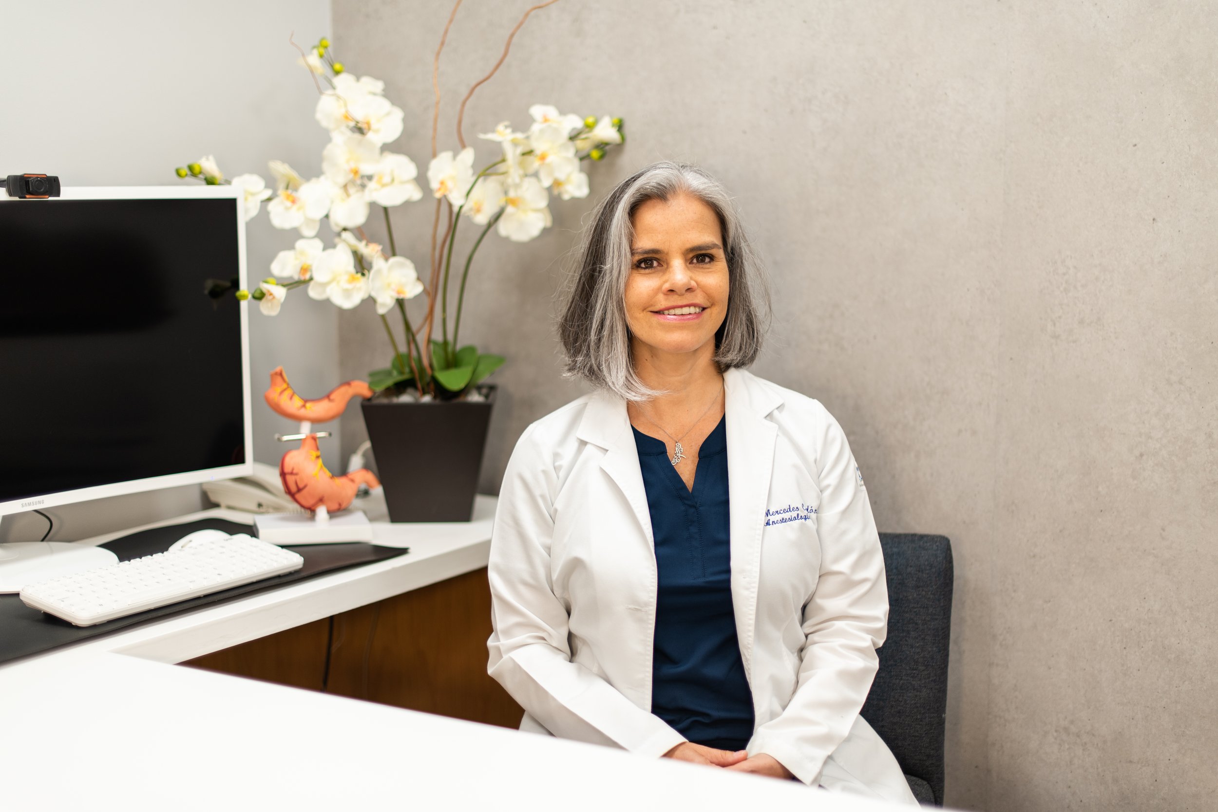 Mujer doctora con bata blanca sentada en consultorio con florero de orquídeas y modelos anatómicos del sistema digestivo en el fondo.