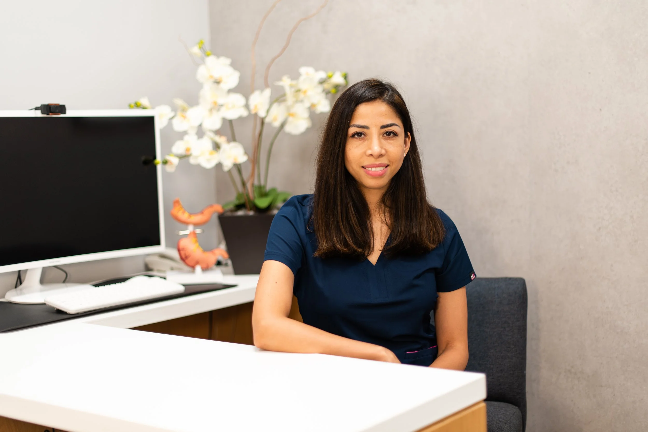 Mujer en uniforme azul sentada en una oficina con planta de orquídeas y modelo de intestinos en el fondo.