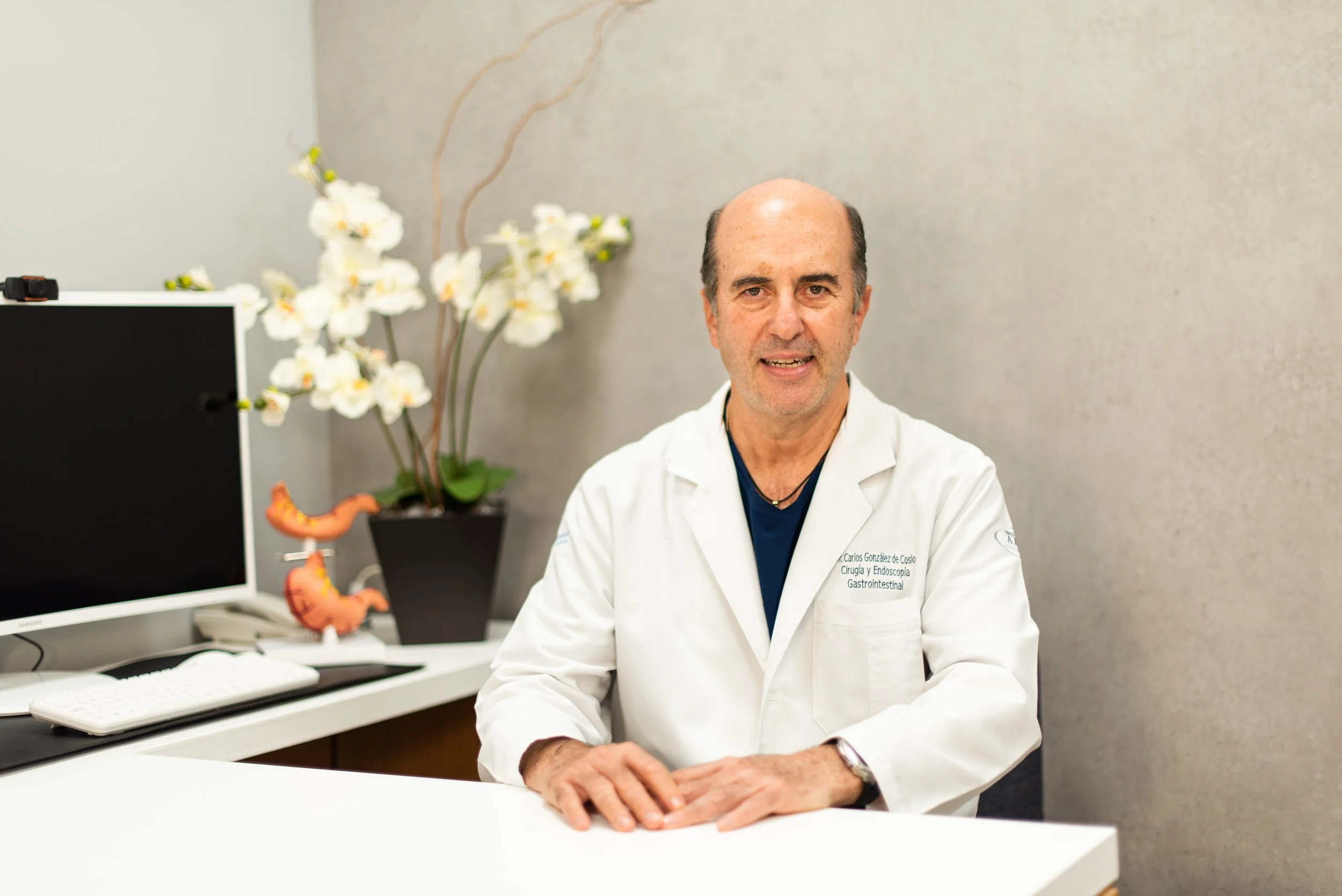 Un médico sentado en un consultorio con plantas y modelos anatómicos de intestinos en el fondo.