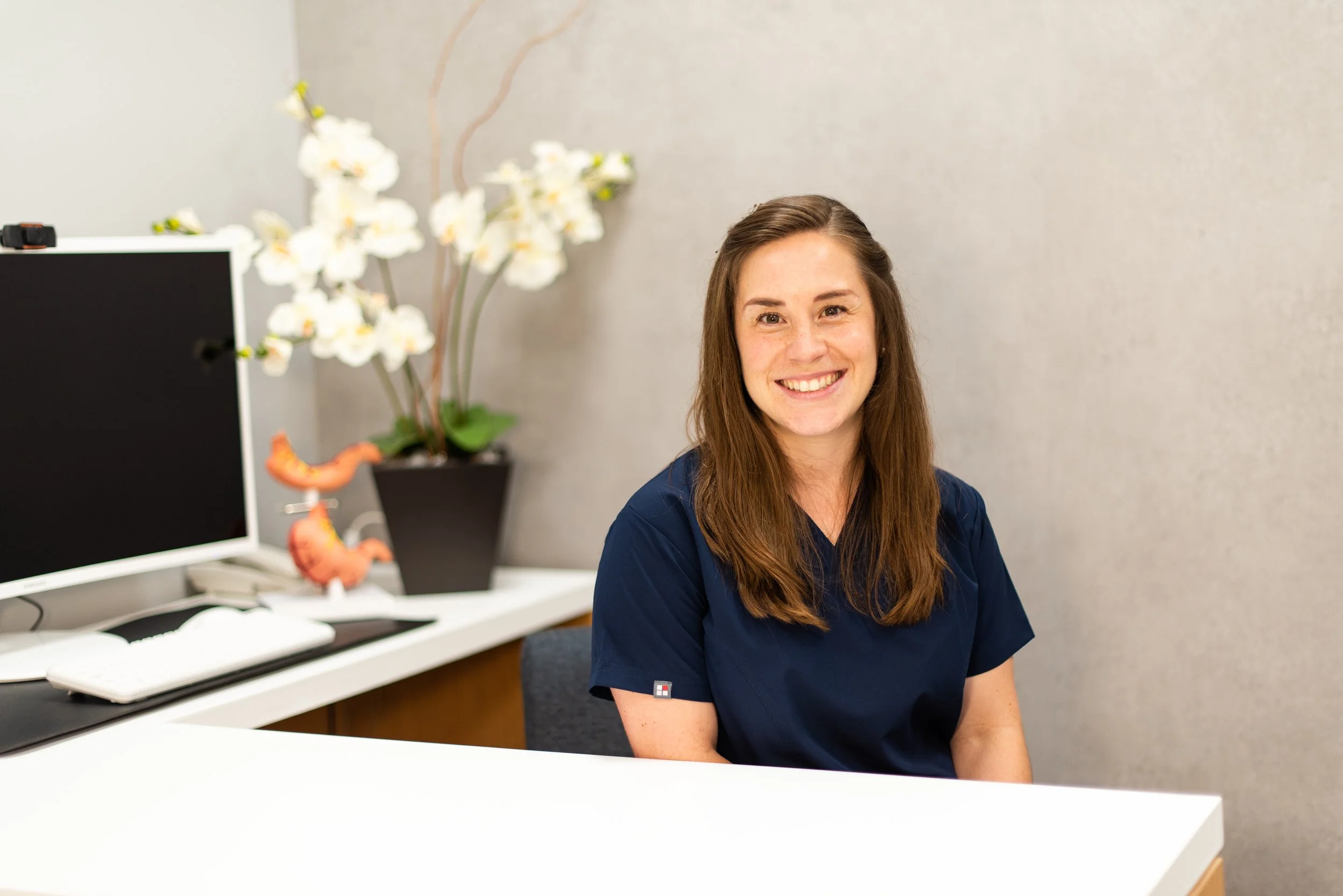 Mujer sonriendo en un consultorio, con una computadora y plantas en el fondo.