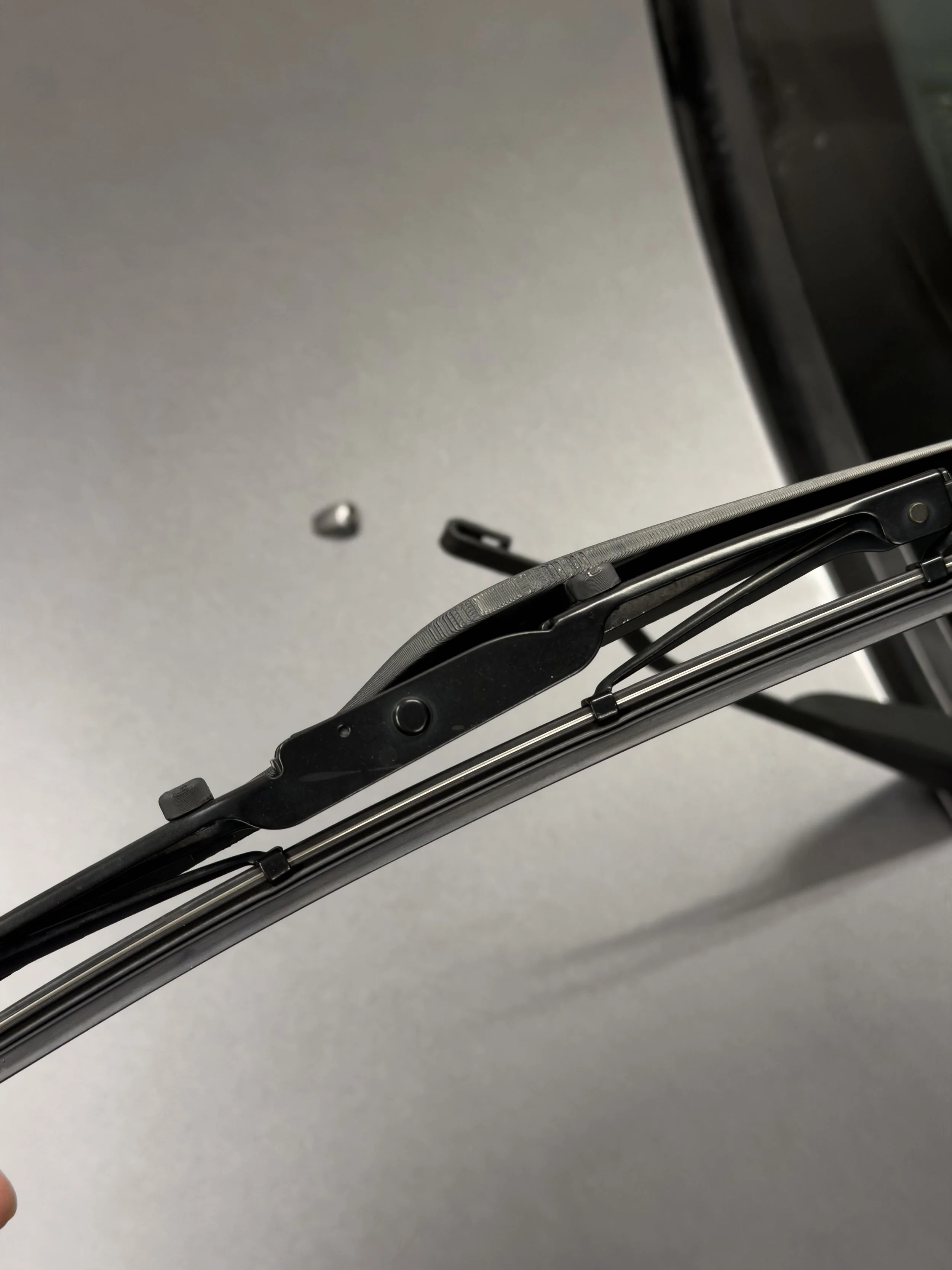 Close-up of windshield wiper arm and blade on a car, with a small metallic object in the background on a gray surface.