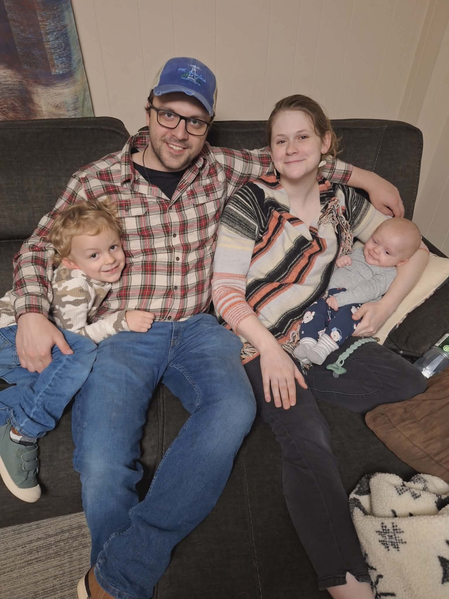 A family sitting on a dark-colored couch, including a man wearing glasses and a blue cap, a woman, a young boy with curly hair, and a baby in a onesie. The boy is leaning on the man's lap, and the woman is holding the baby. They are smiling and appear to be in a cozy living room.