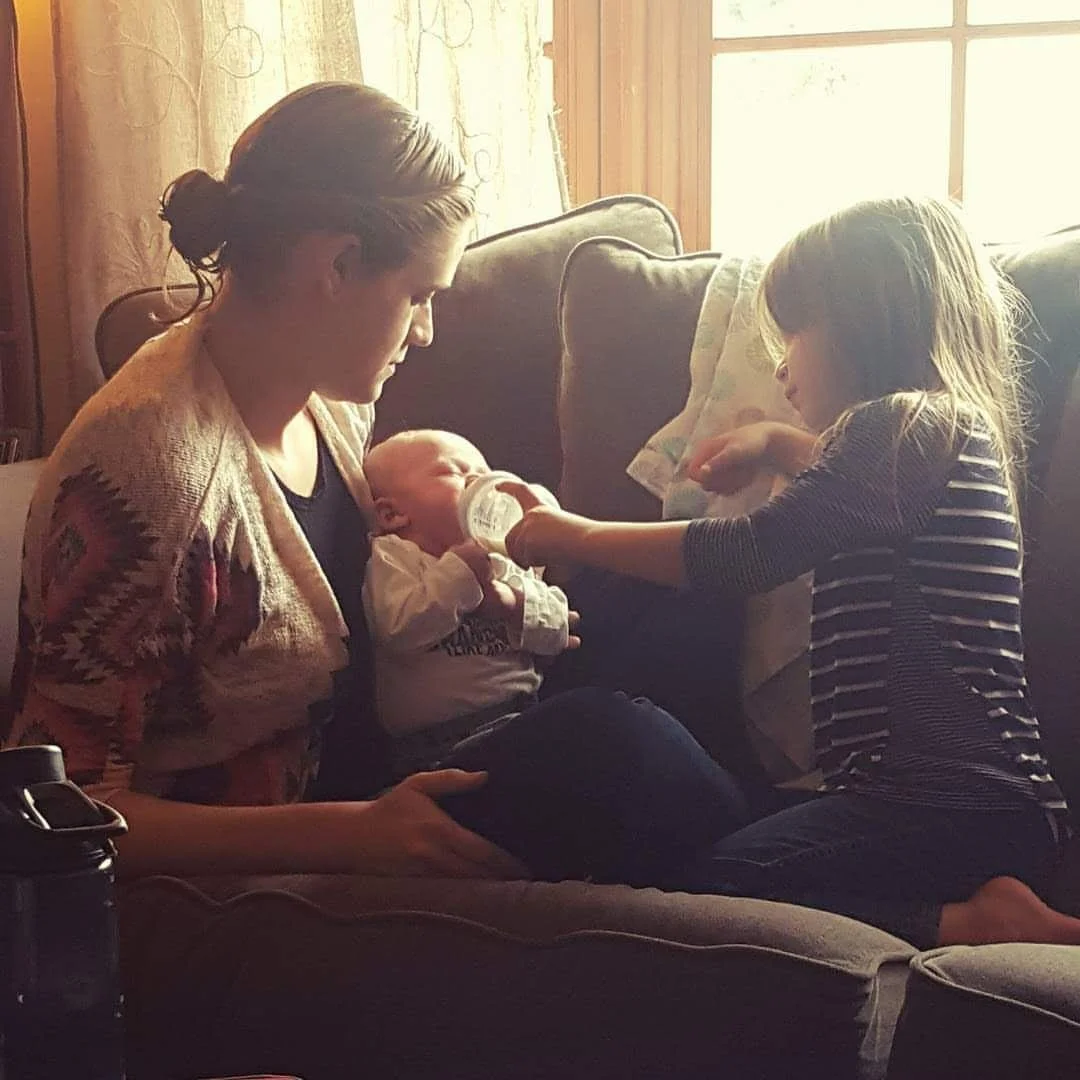A woman, a young girl, and a baby sitting on a sofa in a warm, sunlit living room. The young girl is feeding the baby with a bottle.