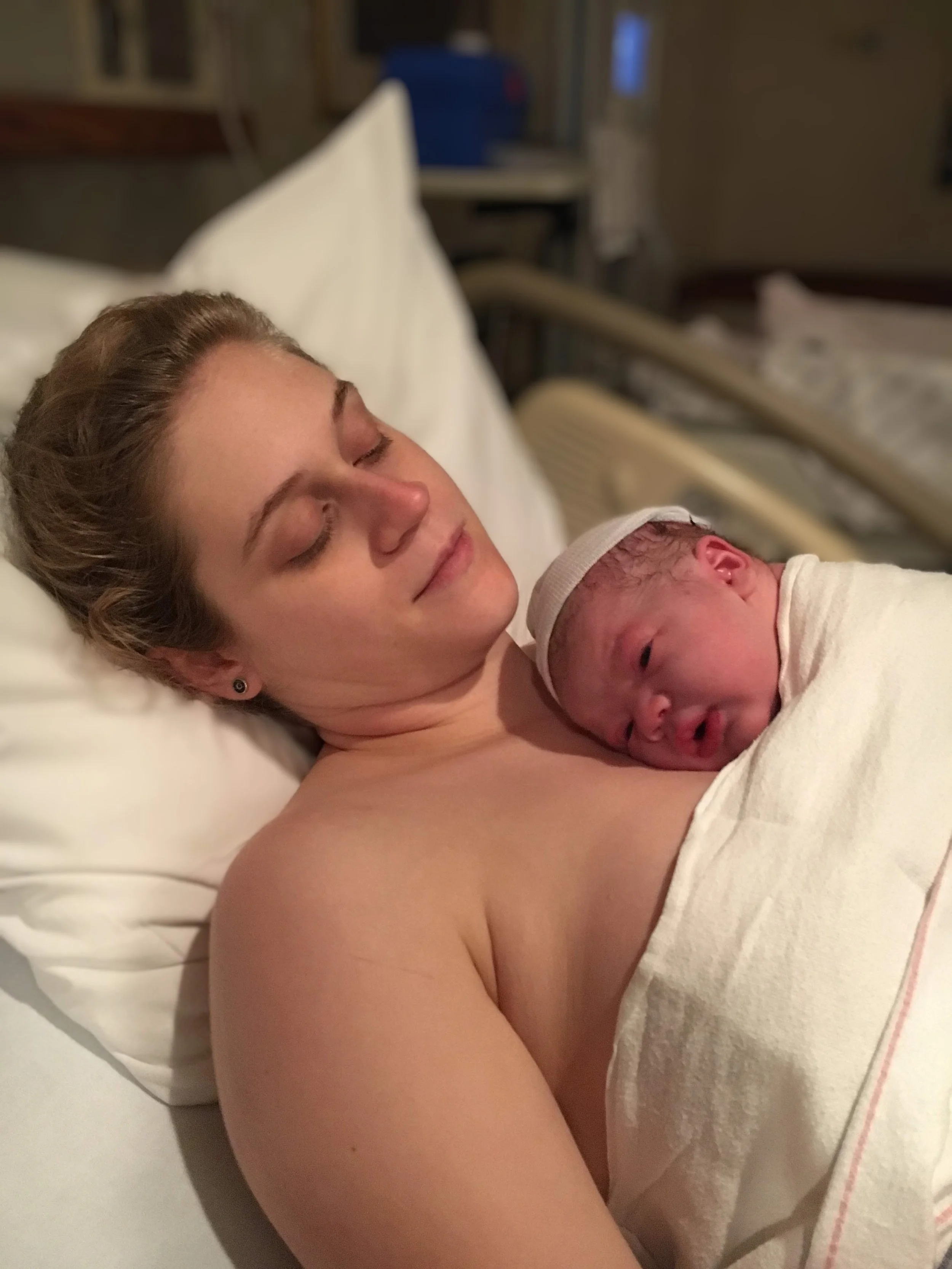 A woman lying in a hospital bed holding a newborn baby close to her chest, both with eyes closed, in a hospital room.