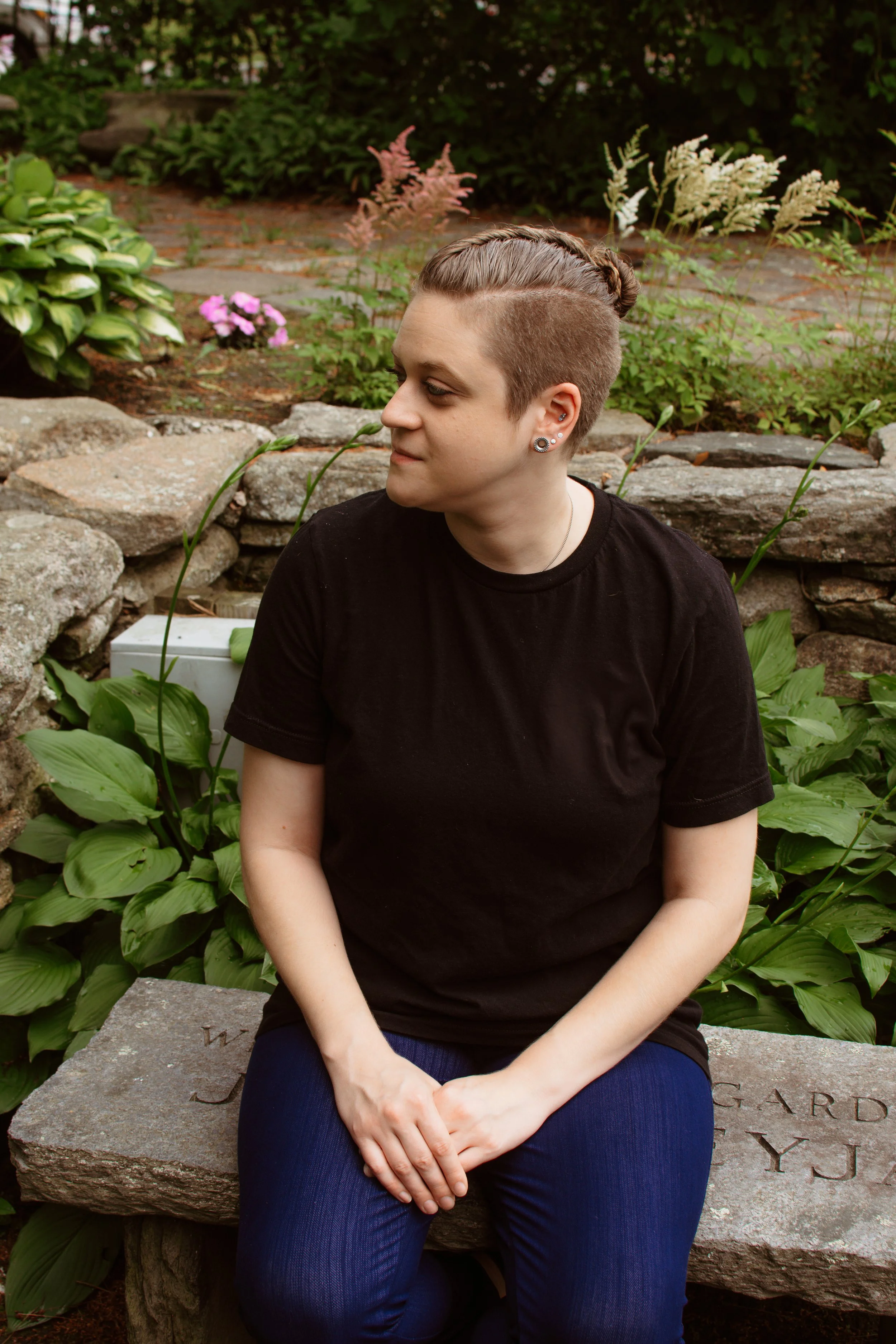 white person sitting down in a place shirt with dark jeans on a stone bench in a garden