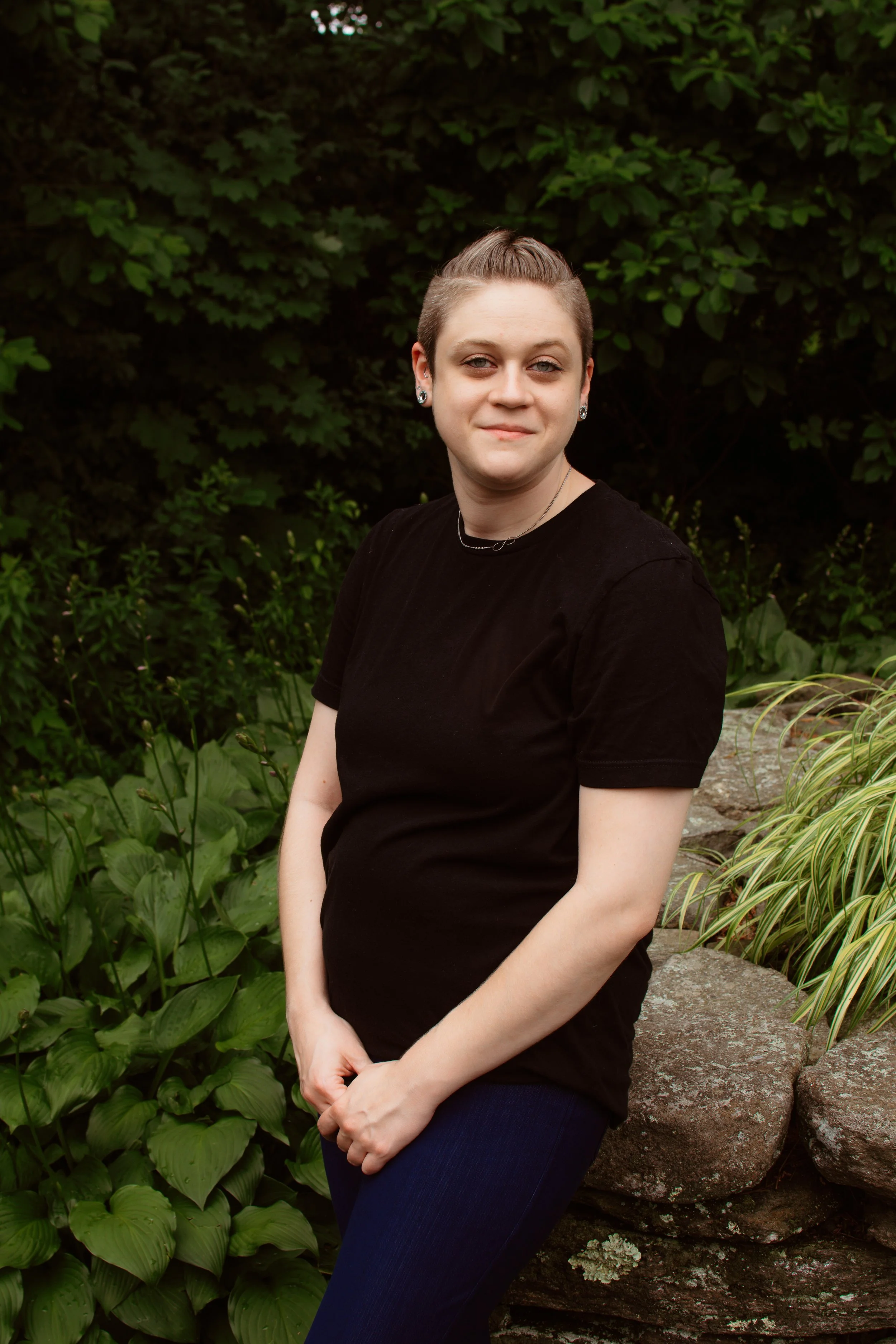 A woman with short hair, wearing a black t-shirt and dark jeans, standing outdoors near green foliage and rocks, looking at the camera.