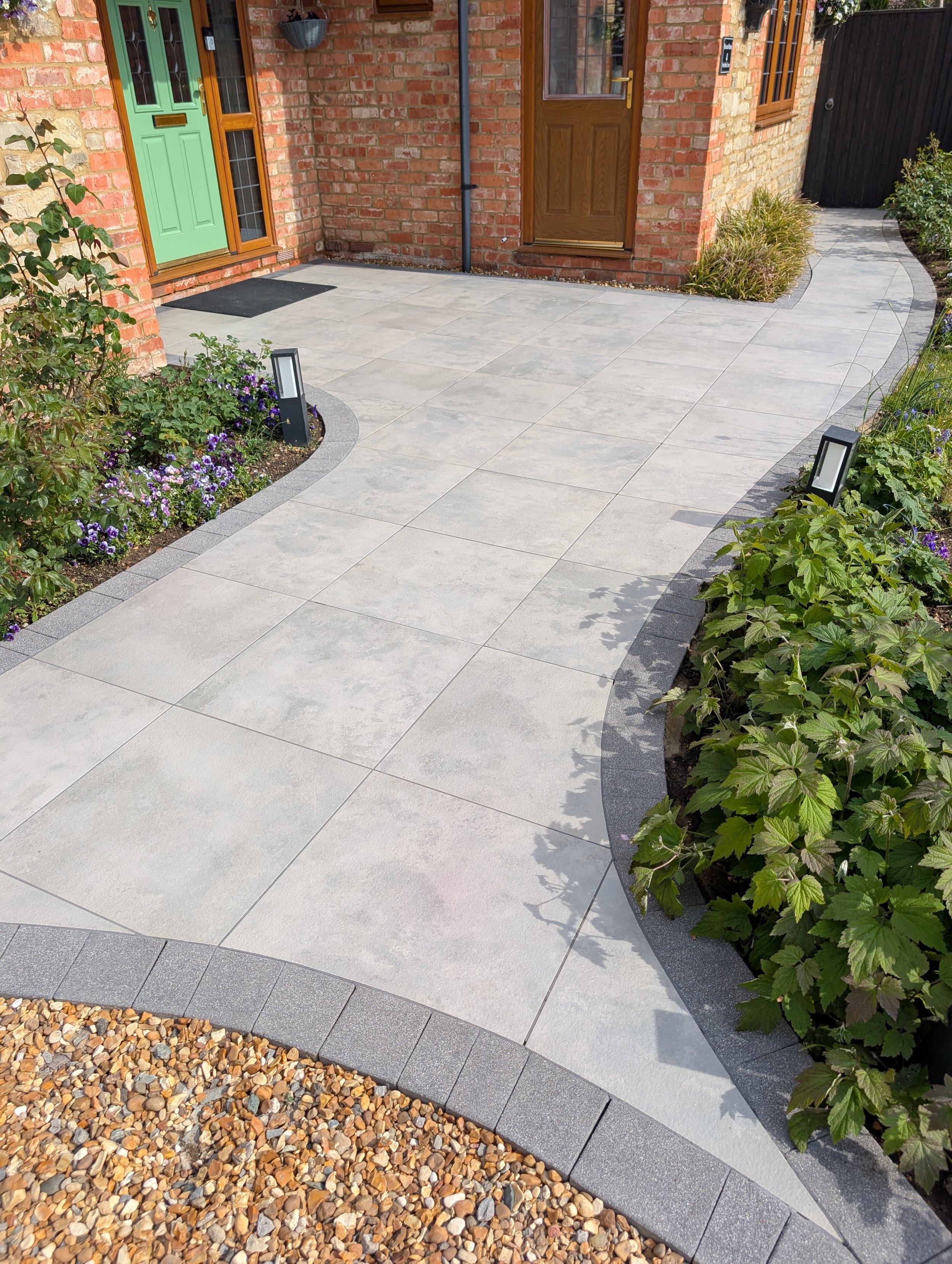 A neatly landscaped front yard with a modern paving pathway, bordered by plants and small garden beds, leading to a brick house with a bright green front door and a wooden front door beside it. The yard includes garden lights, a gravel area, and a bl