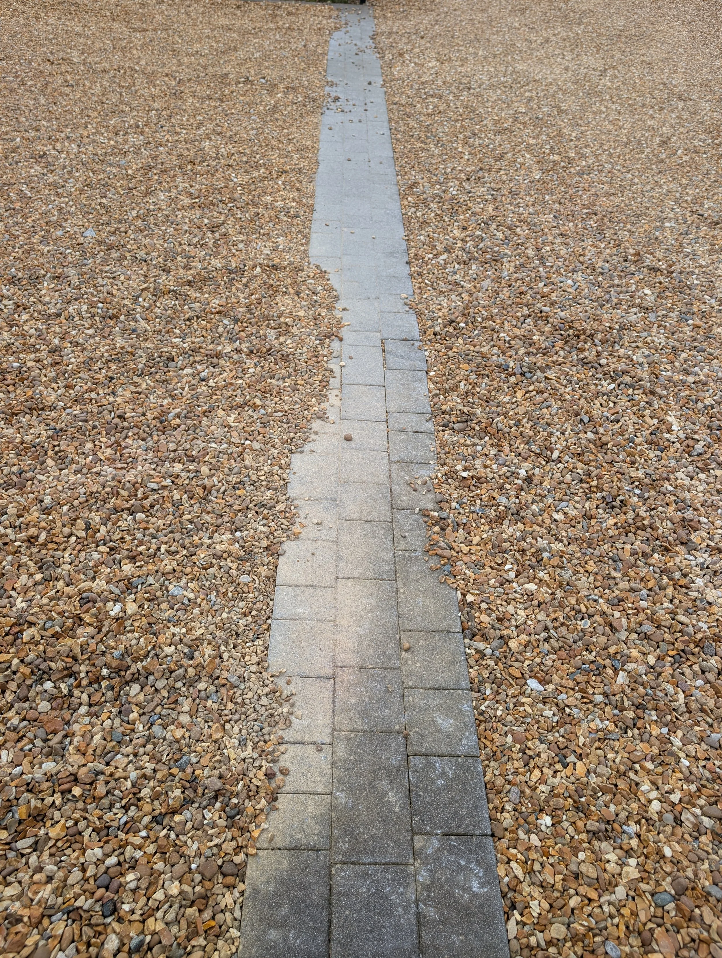 A dirty and concrete covered narrow paved pathway made of rectangular concrete block paving bricks, surrounded by small multicoloured gravel stones.
