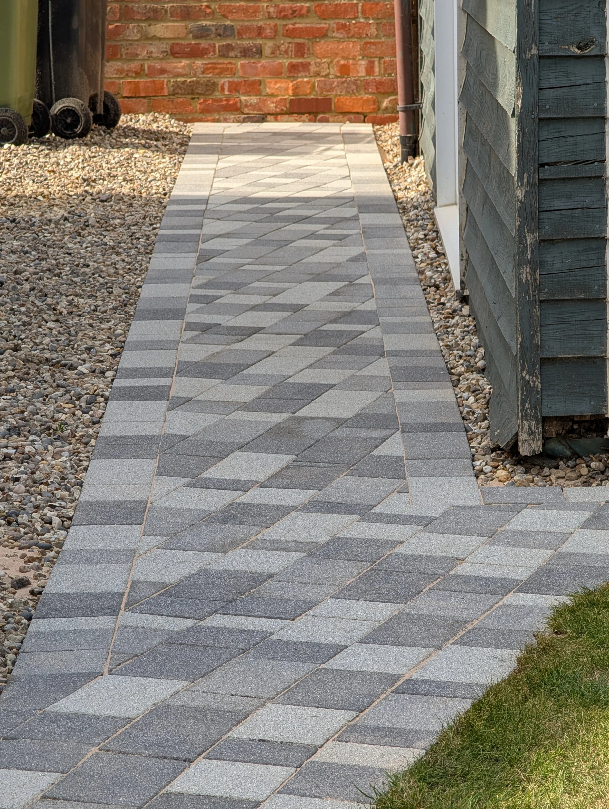 A freshly cleaned, chemically treated and resanded narrow paved walkway made of rectangular grey concrete block paving stones, next to a gravel area and a house with blue siding.