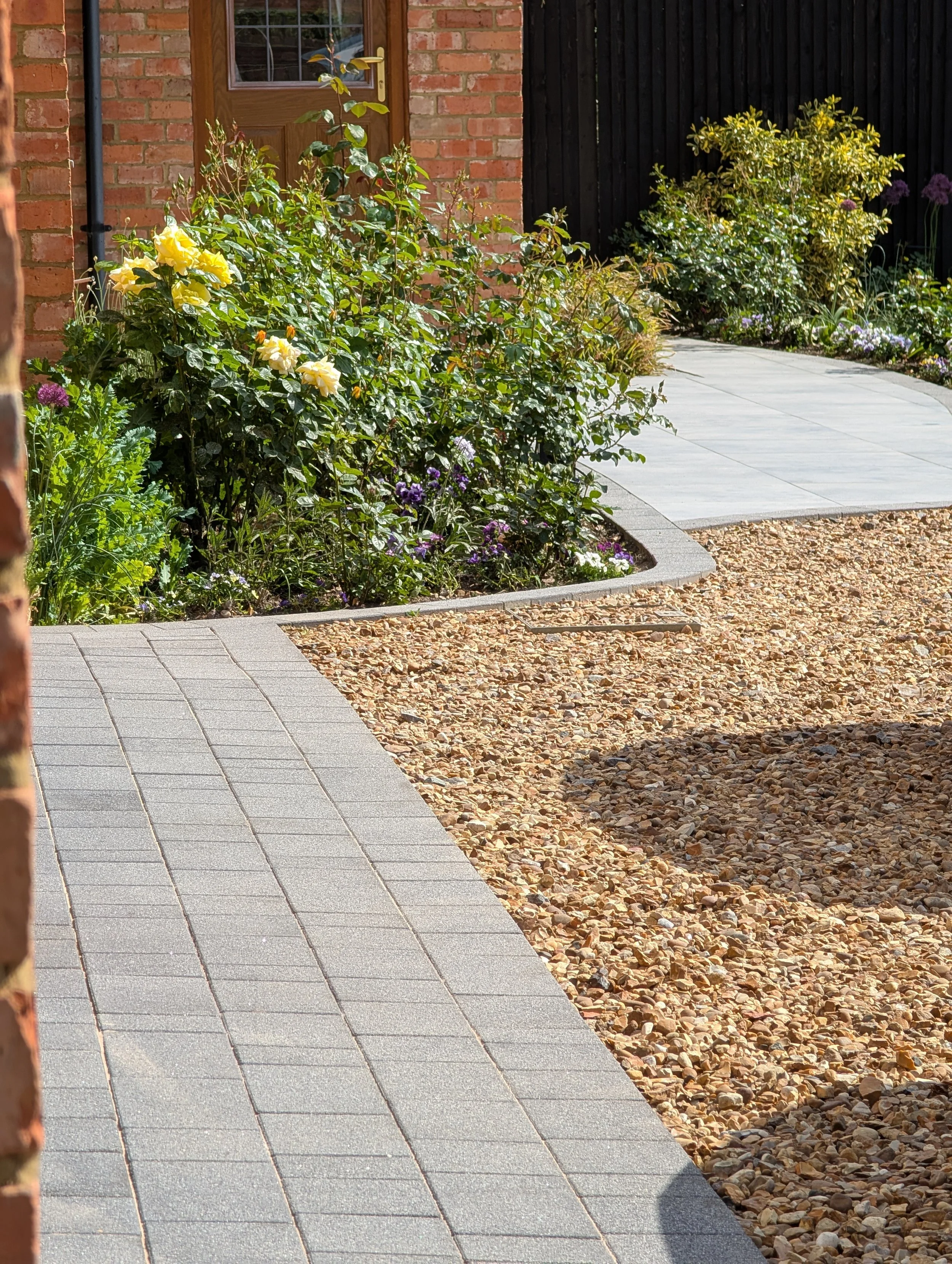 A garden pathway with stone tiles, surrounded by flower beds with yellow, purple, and white flowers, next to a brick house with a wooden door.