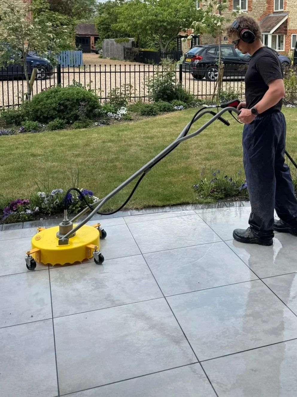 Person using a floor cleaning machine on a tiled patio outdoors, with a garden and houses in the background.
