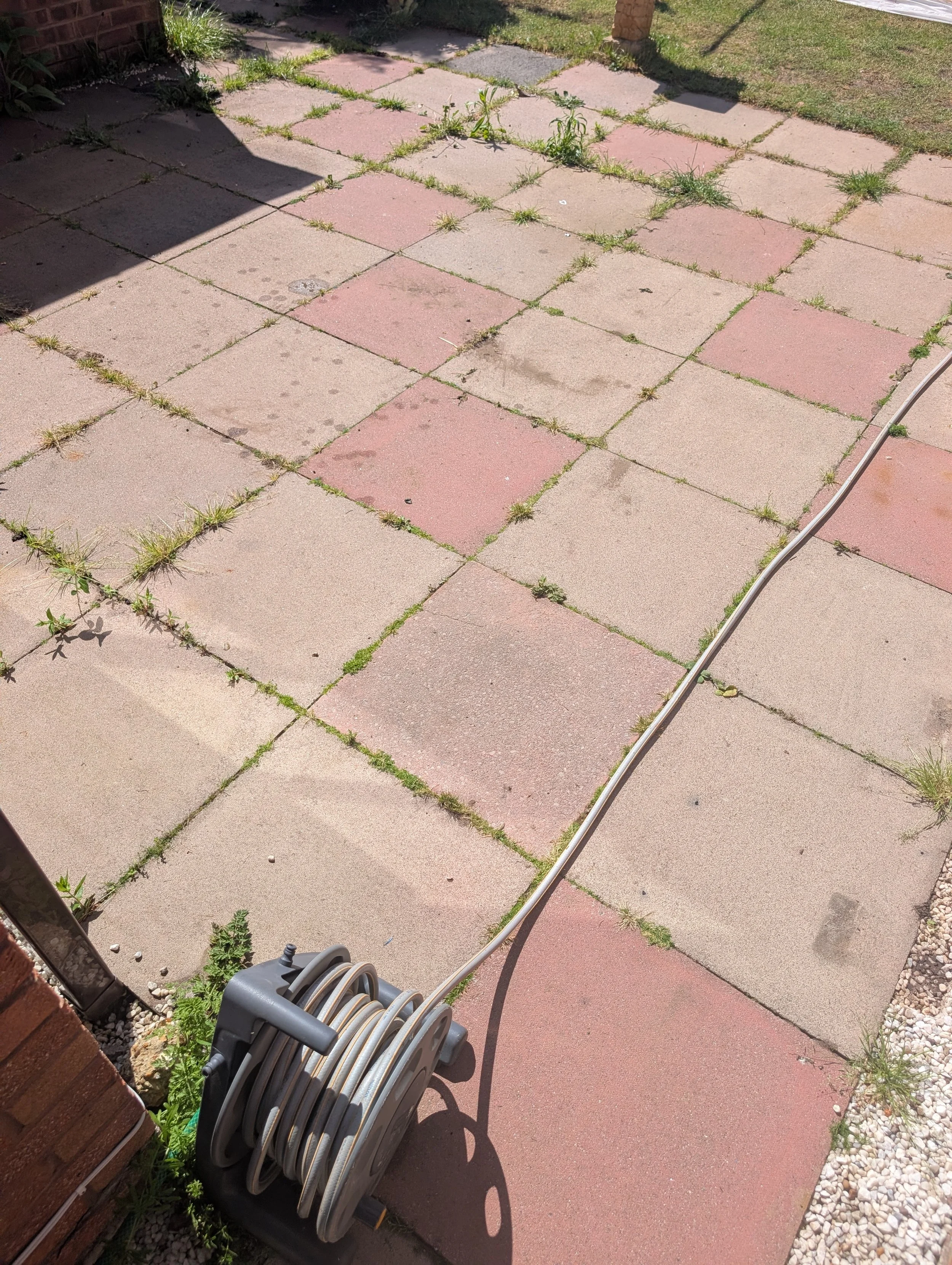 A backyard patio with large square paving stones, some grass and weeds growing between the stones, a garden hose reel near a brick wall, and a garden garden on the upper right side.