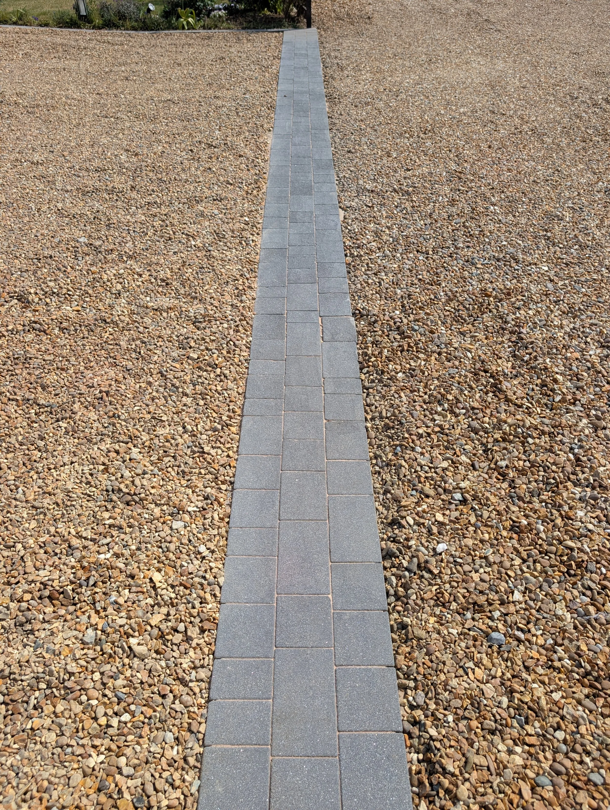 A freshly cleaned, chemically treated and re-sanded narrow paved pathway made of rectangular concrete block paving bricks, surrounded by small multicoloured gravel stones.