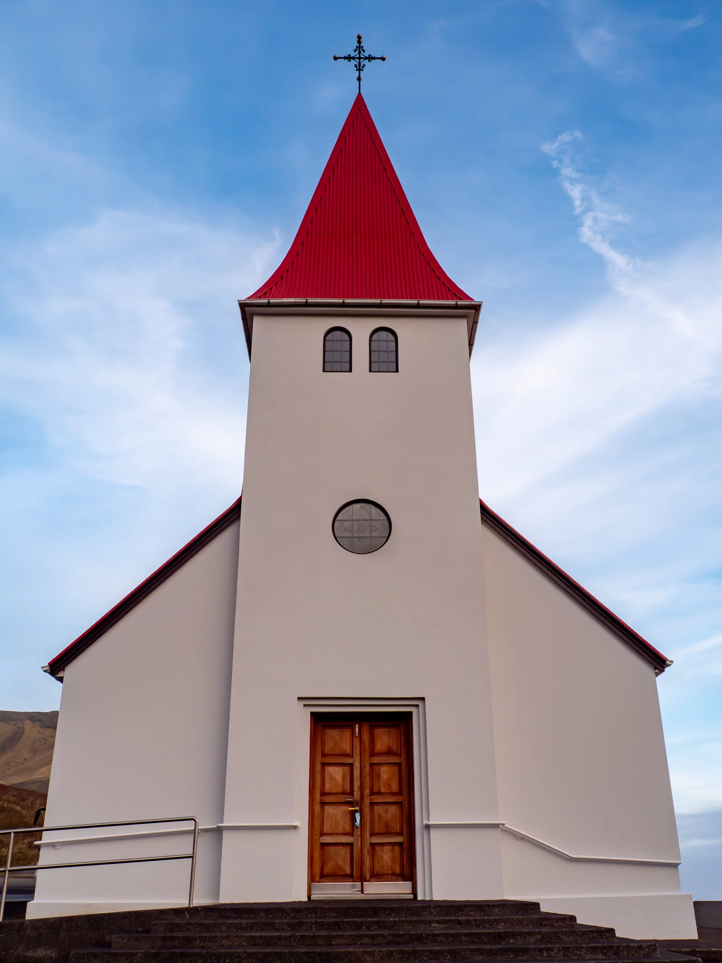 A white church with a steep red roof and circular clock window, with a wooden double door entrance, set against a blue sky with some clouds.