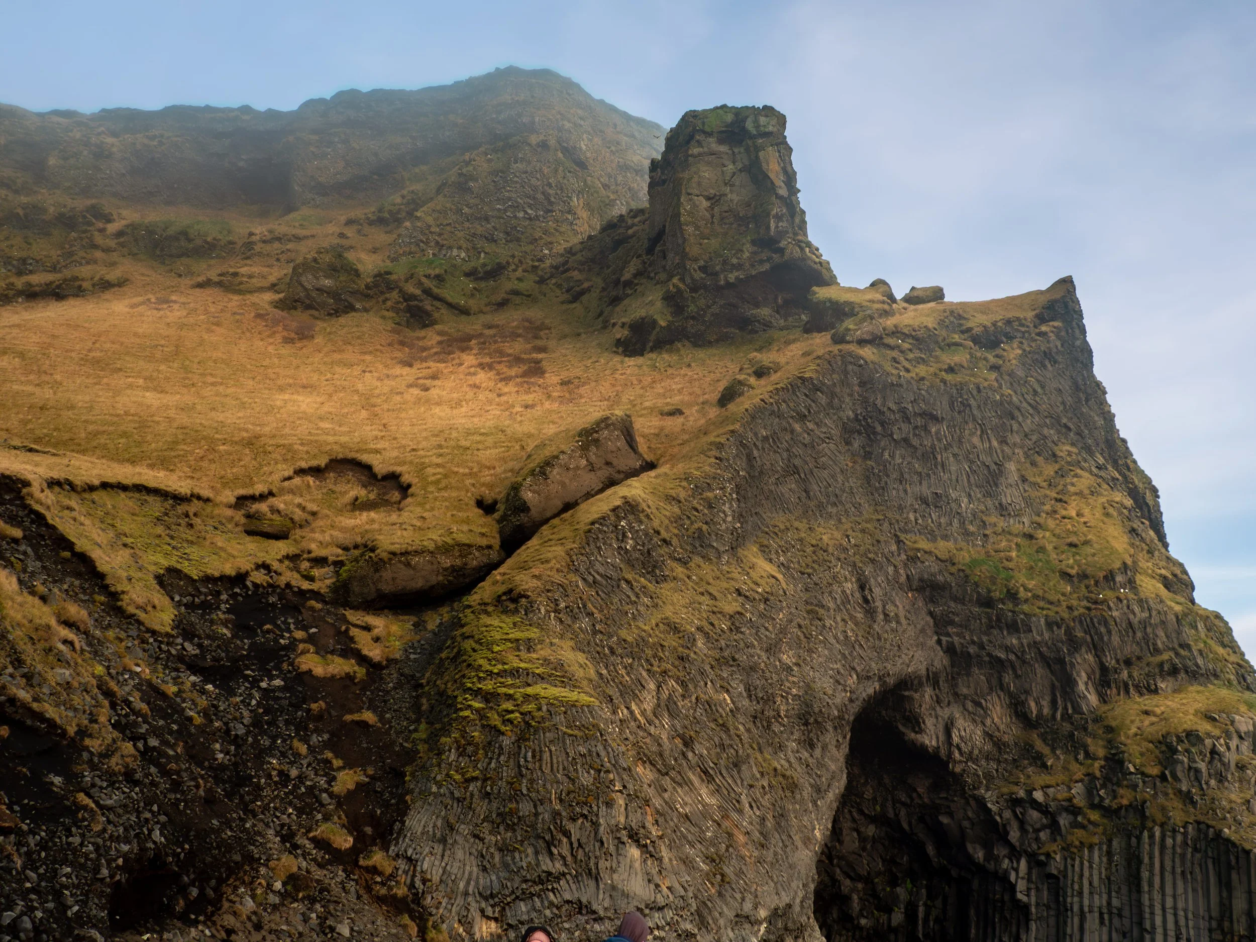 A rugged mountain landscape with steep cliffs, grassy slopes, and a natural arch formation in the foreground.