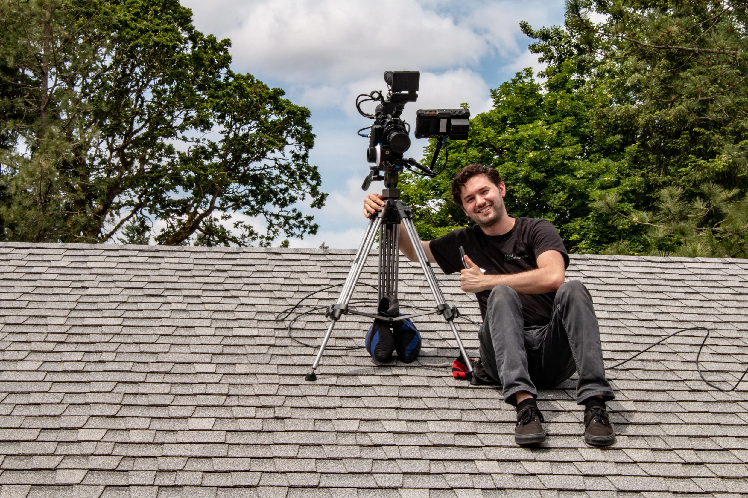 A young man sitting on a roof with a camera mounted on a tripod, smiling and giving a thumbs-up.